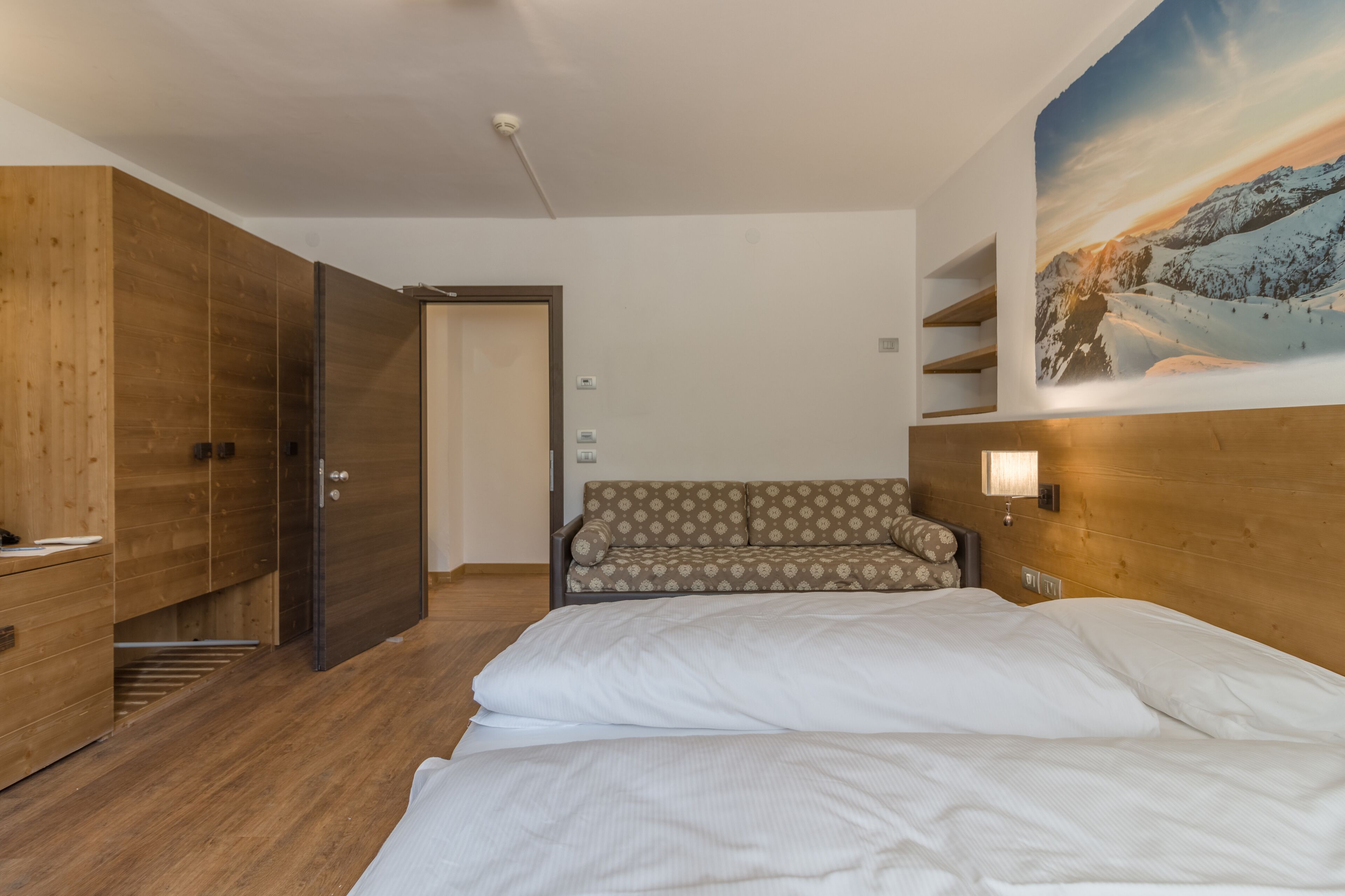 A hotel room with a wooden wardrobe, a sofa, a bed, and a painting of a snowy mountain landscape.