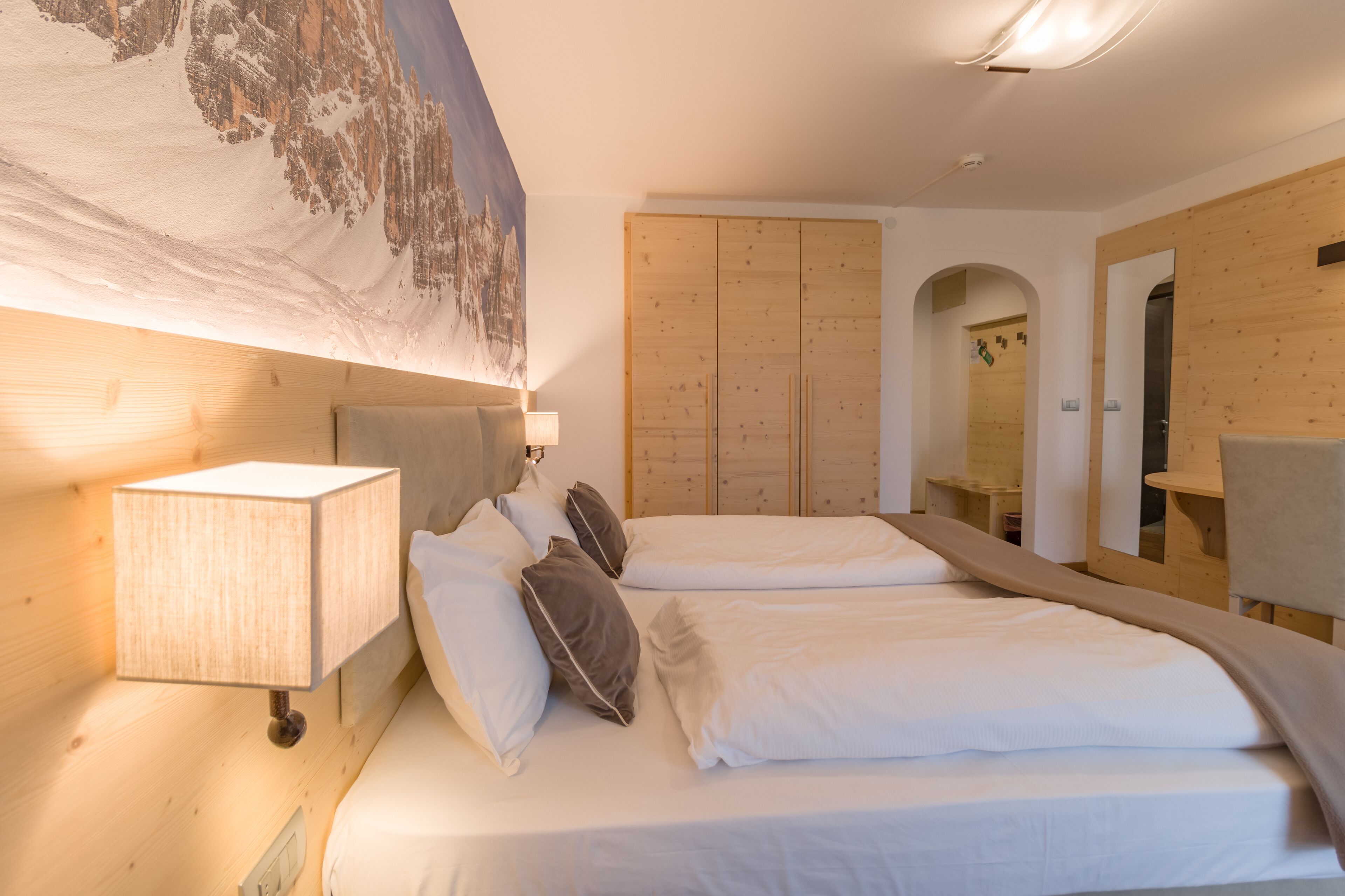 A hotel room with two beds, a wooden wardrobe, a mirror, and a painting of a mountain landscape.