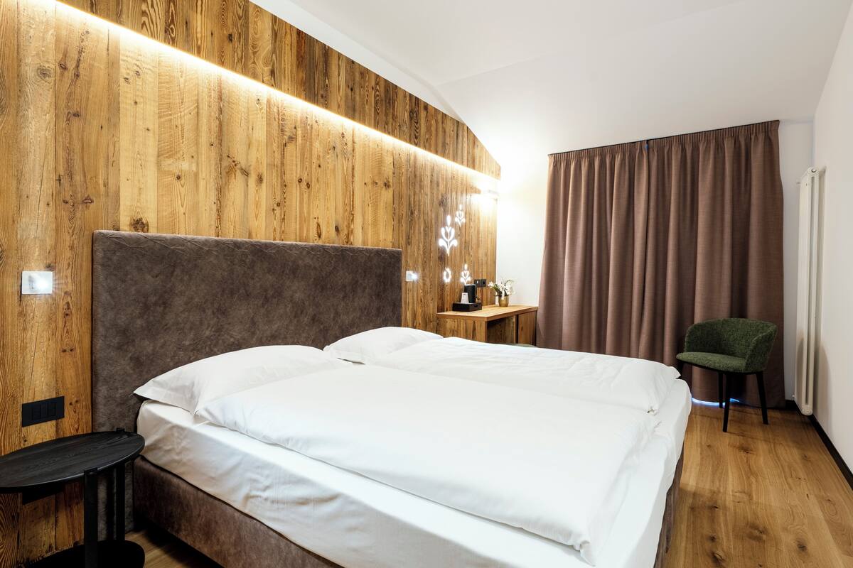 Une chambre d’hôtel avec une tête de lit en bois, un lit avec du linge de lit blanc, une table de chevet, une chaise verte et une fenêtre avec des rideaux.