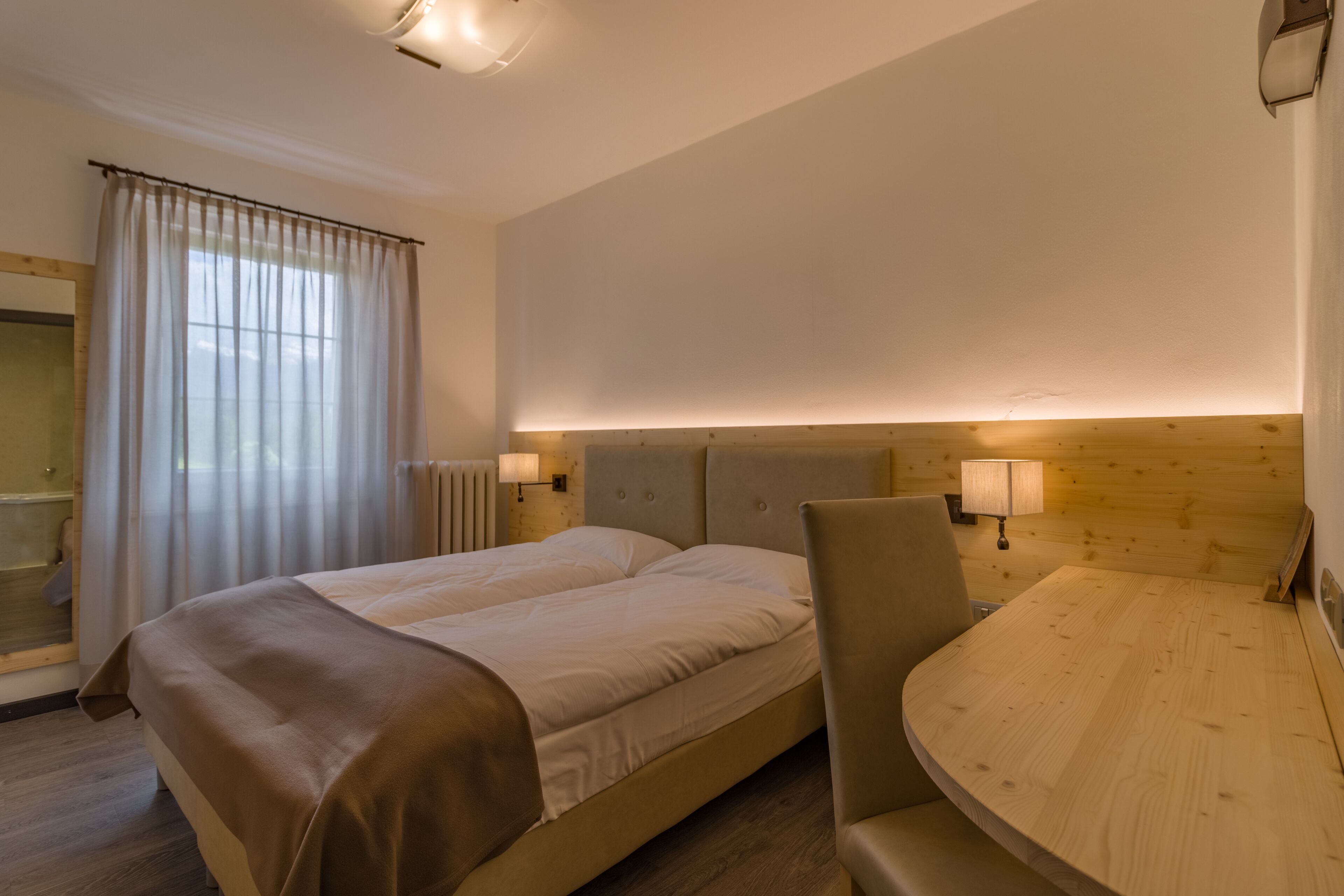 Habitación de hotel con cama doble, cabecera de madera, una mesita pequeña y una silla. Se ve una ventana con cortinas y una lámpara de pared.