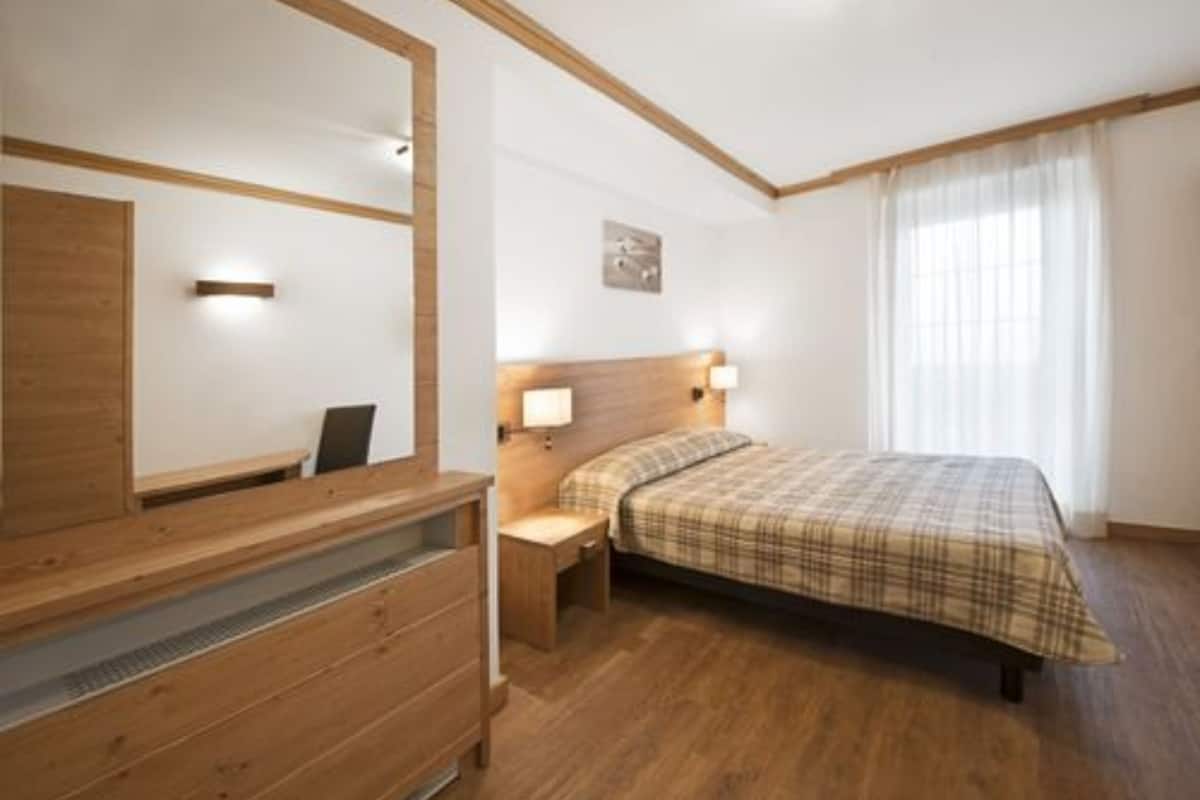 Une chambre avec une commode en bois, un lit recouvert d’une couverture à carreaux et deux lampes de chevet.