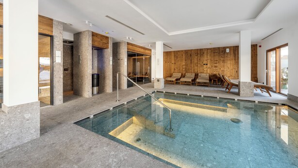Ein überdachter Schwimmbereich mit Blick auf das Becken, einer Holzwand und einer Lounge-Ecke mit Liegestühlen.