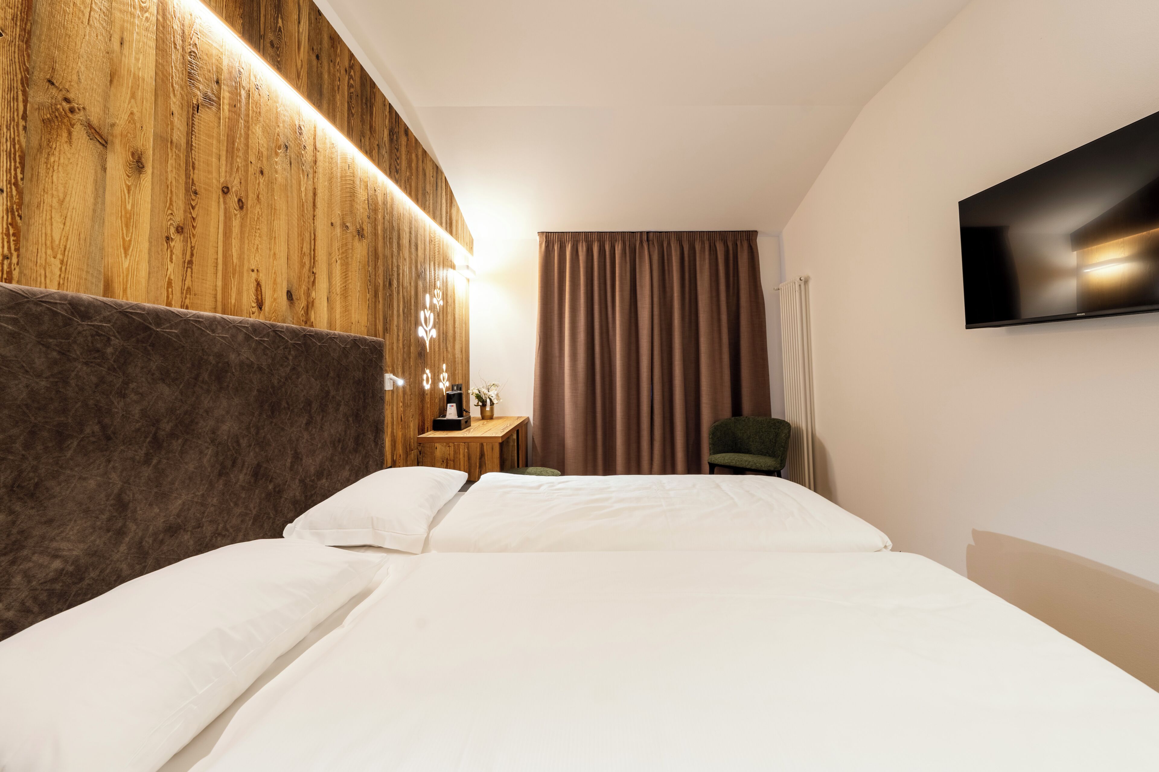 Habitación de hotel con cabecera de madera, televisor de pantalla plana, cama con ropa blanca, mesita de madera pequeña y una silla verde.