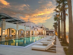 A pool area with lounge chairs and a covered seating area, surrounded by palm trees and a building with arches. - Grecotel Plaza Beach House (Rethymno)