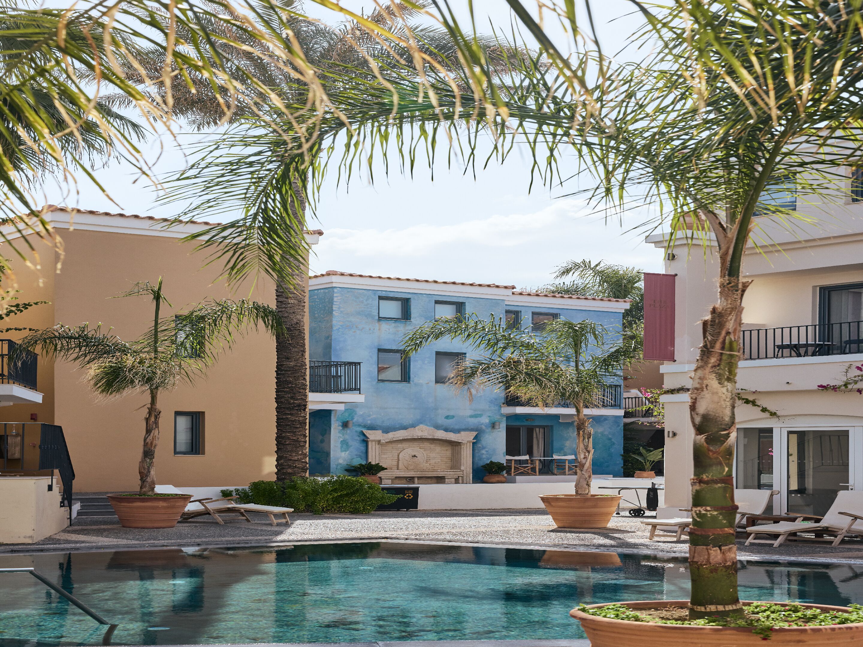 Un área de piscina con palmeras, un edificio azul y un edificio color arena.