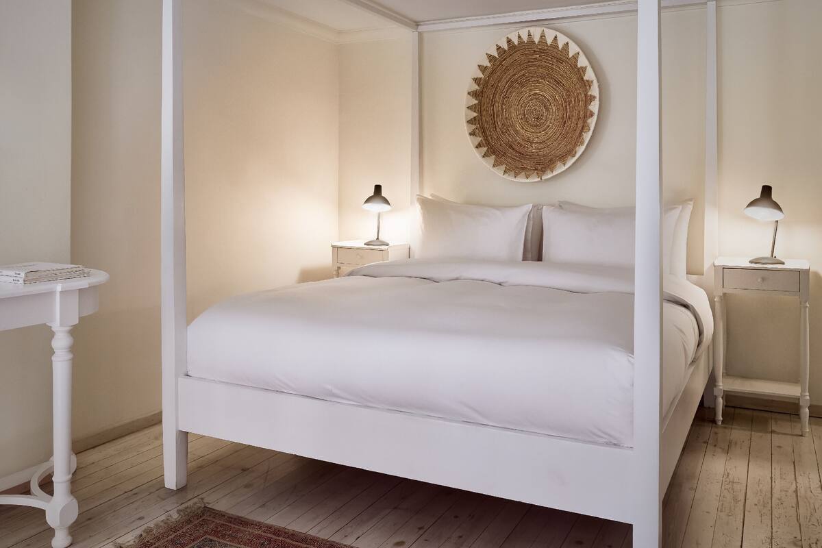 A four-poster bed with white bedding, two bedside tables with lamps, a round woven wall art piece, and a small white table with a rug.