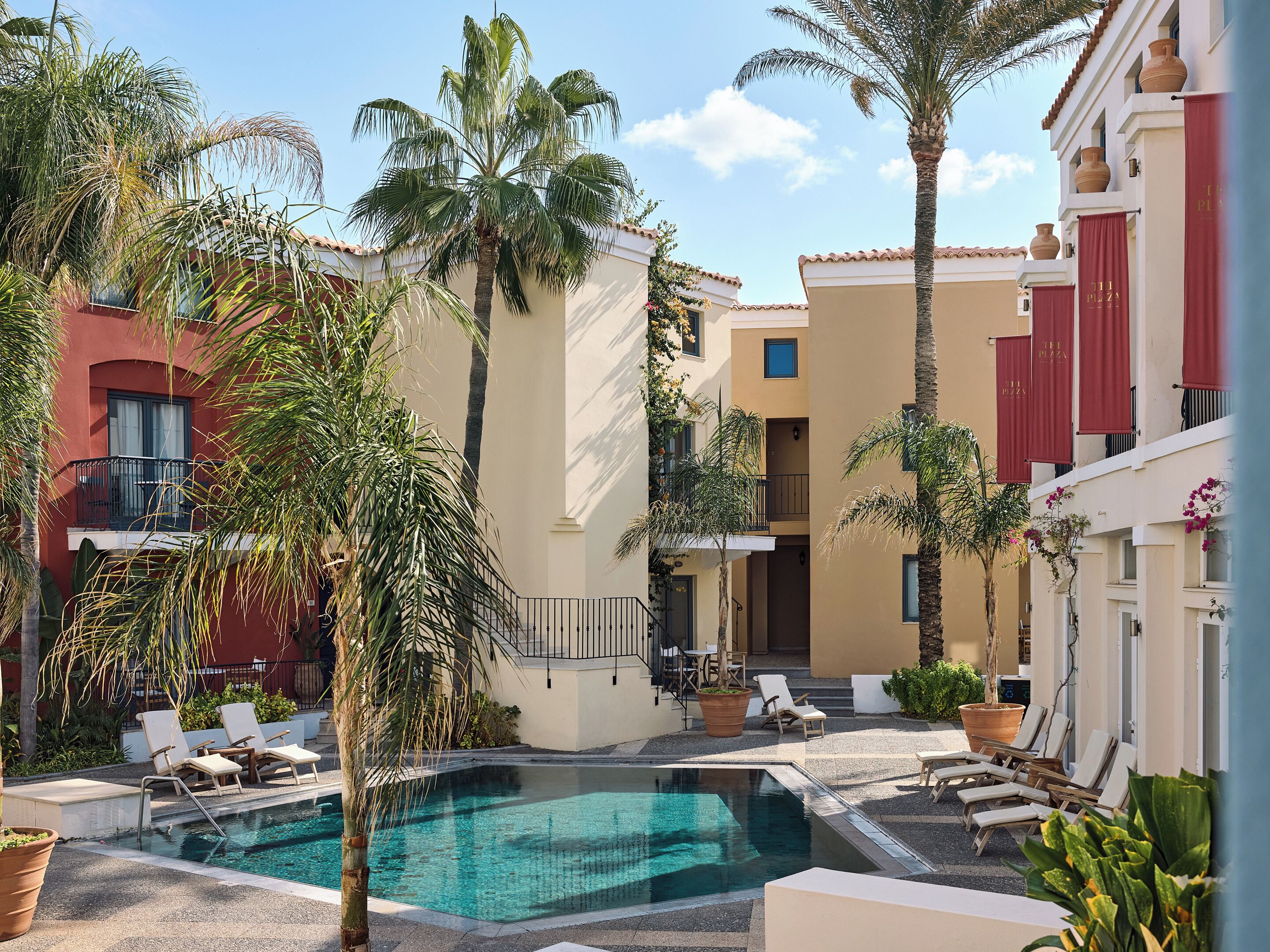A pool area with lounge chairs, palm trees, and a building with red awnings.