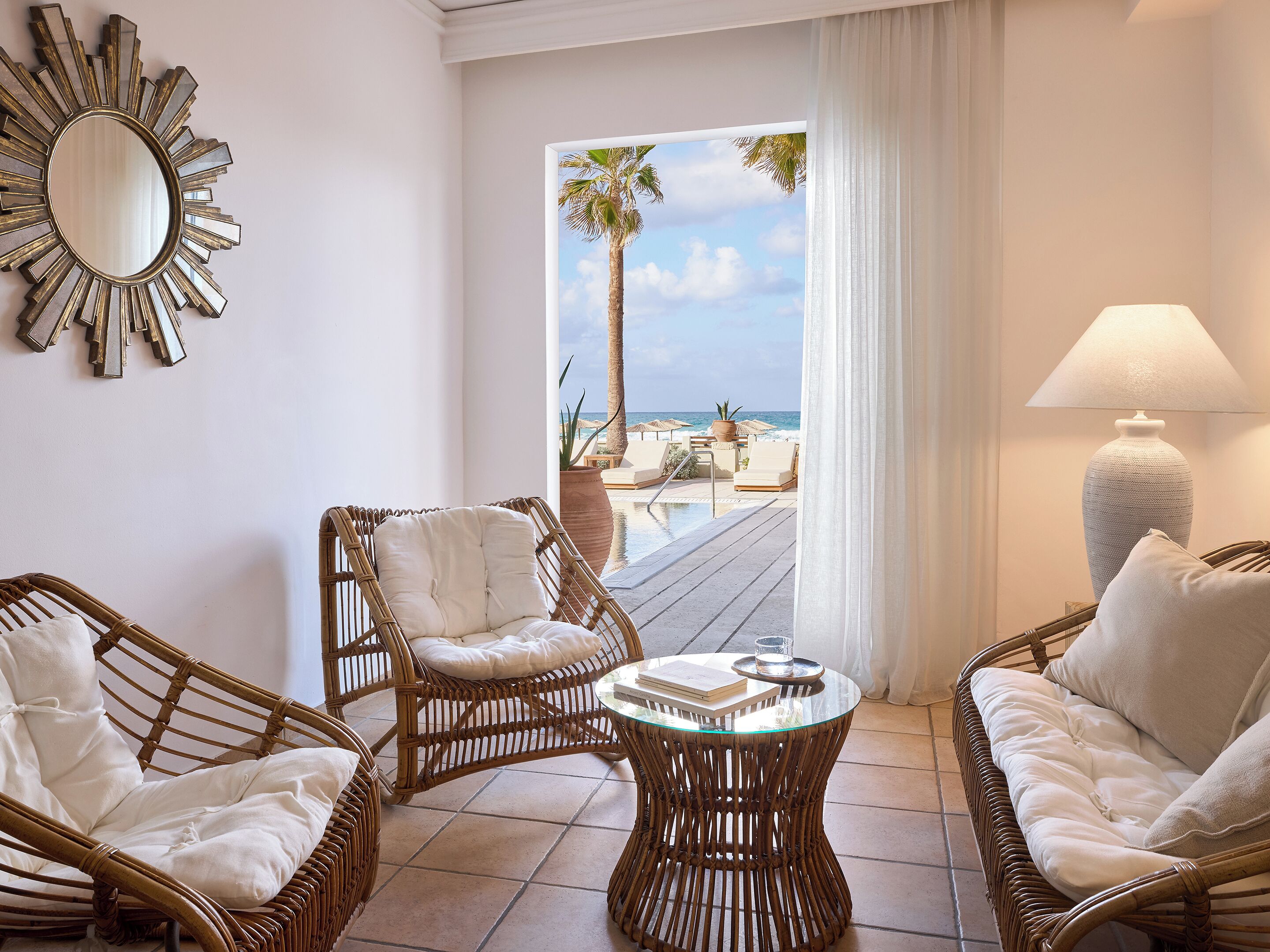A room with wicker furniture, a round table, a large mirror, and a view of the beach.
