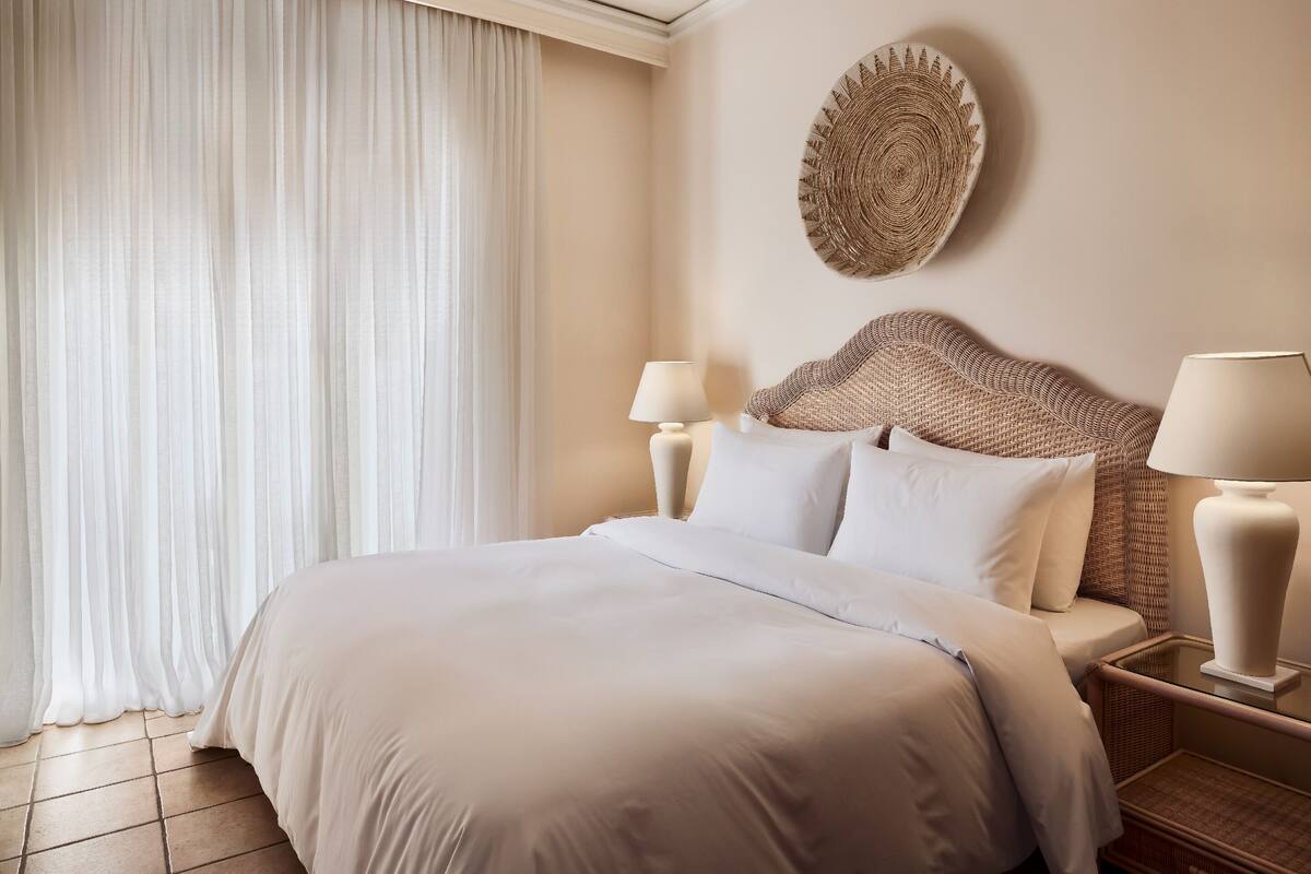 A hotel room with a large bed, two bedside lamps, a woven headboard, and a decorative wall piece.