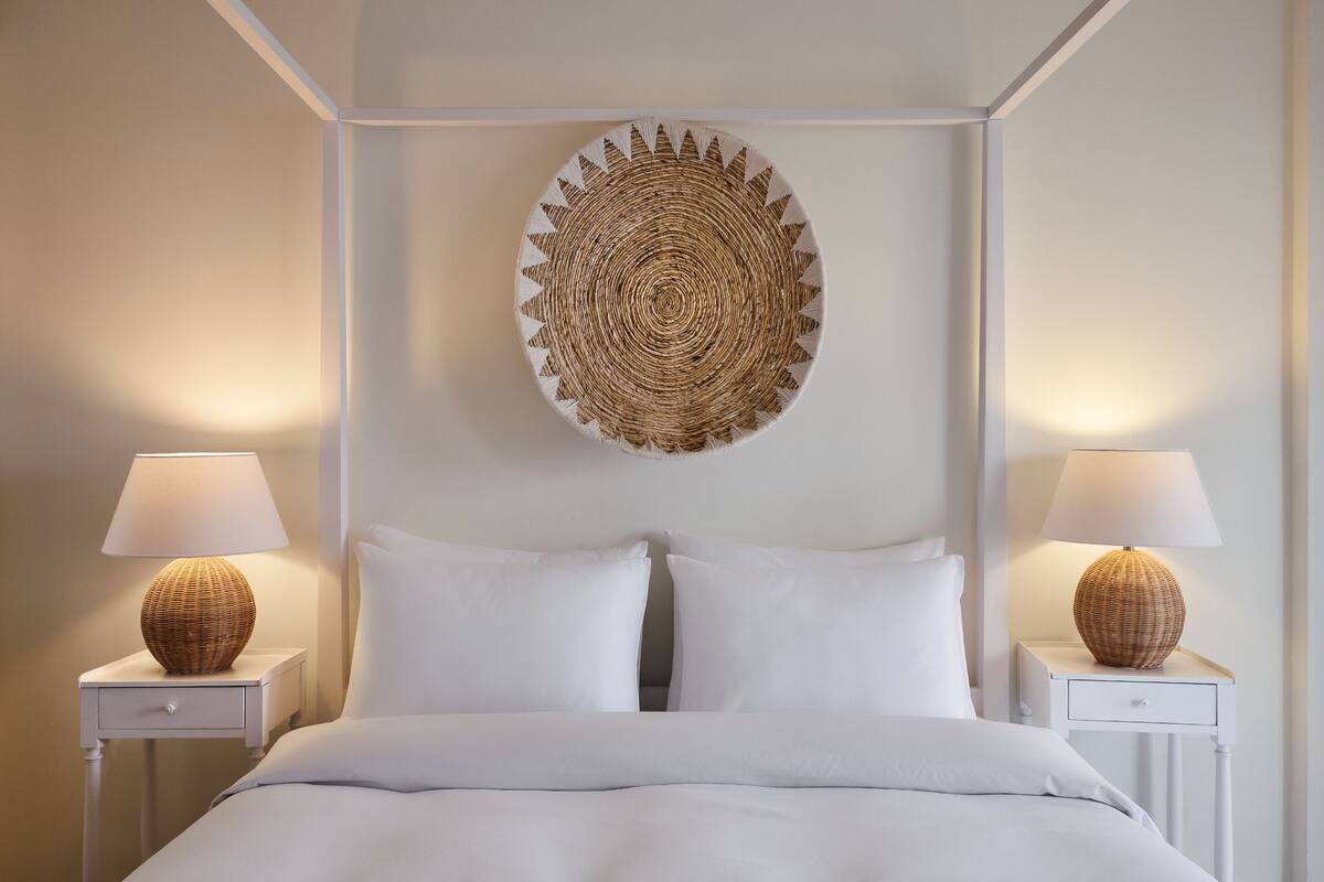 A neatly made bed with white linens, two bedside lamps with woven bases, and a circular woven wall hanging above.