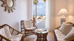 A room with wicker furniture, a round glass table, a large mirror, and a view of a beach and palm trees through a window. - Grecotel Plaza Beach House (Rethymno)