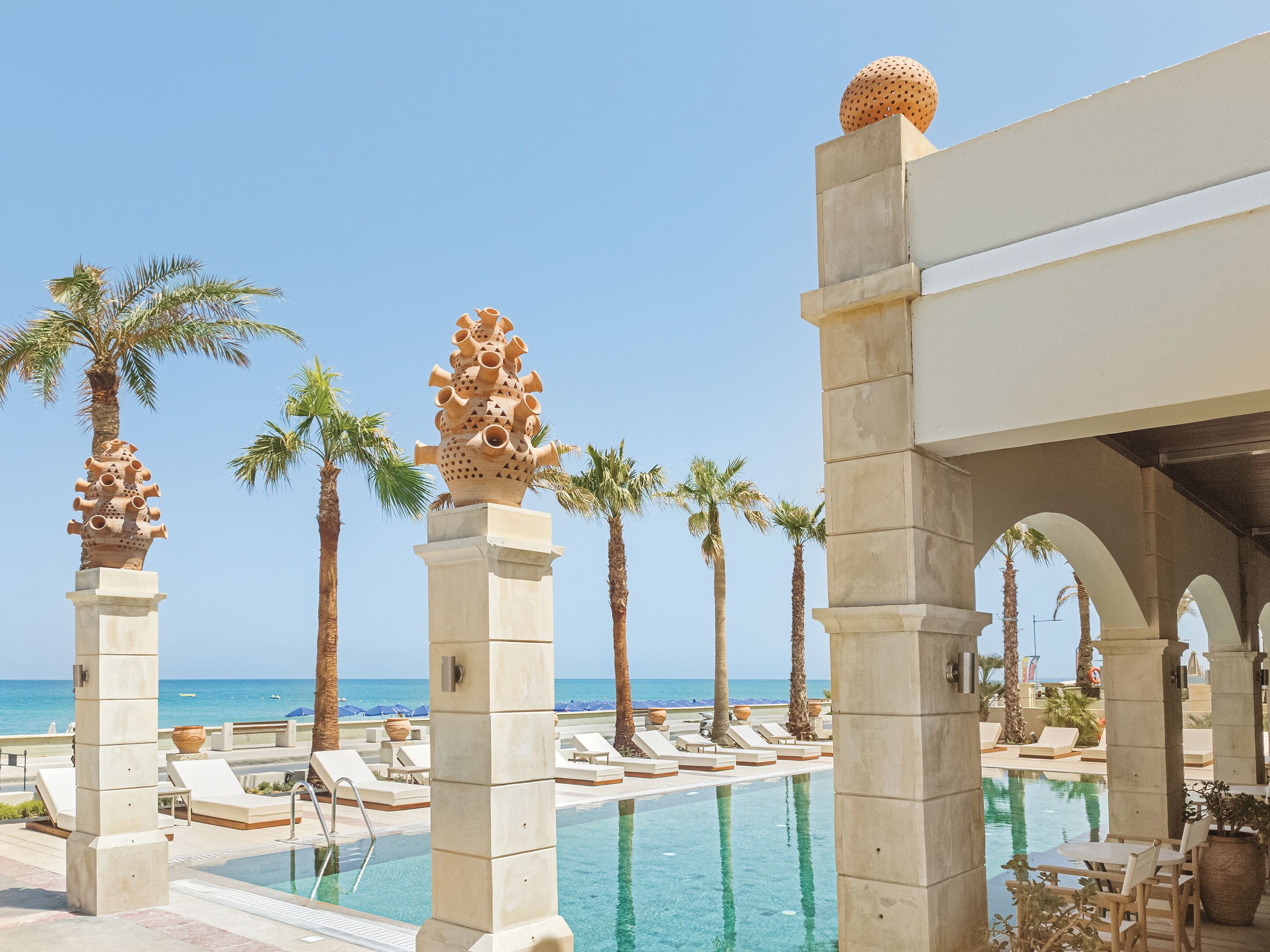 A poolside area with lounge chairs, palm trees, and a view of the ocean.