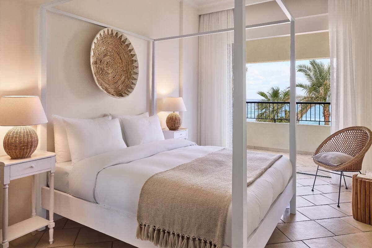 A four-poster bed with white bedding, a woven wall hanging, and a view of the sea through a balcony.