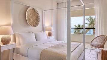 Una cama con dosel con ropa de cama blanca, un tapiz tejido y vista al mar desde un balcón.