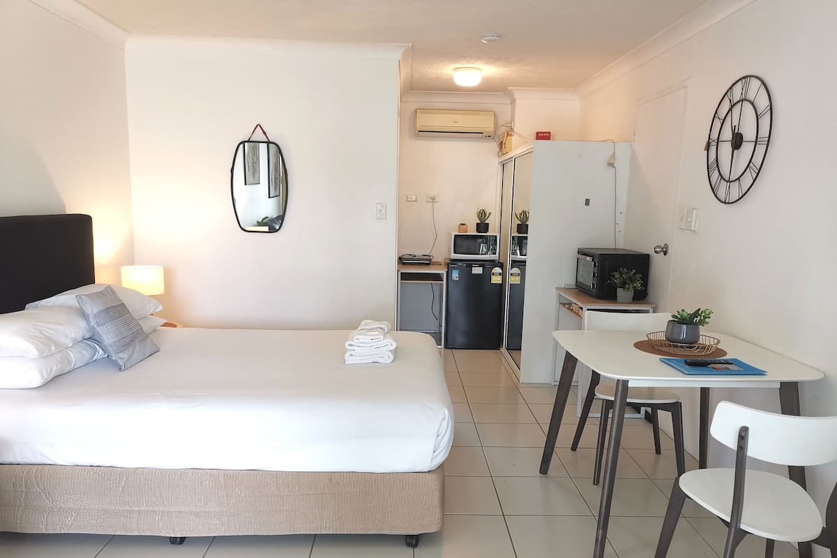 A hotel room with a bed, bedside table, lamp, and a kitchenette area with a microwave, refrigerator, and small potted plants.