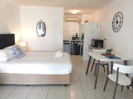 Habitación de hotel con cama, mesita de noche, lámpara y una kitchenette que incluye microondas, refrigerador y pequeñas macetas.