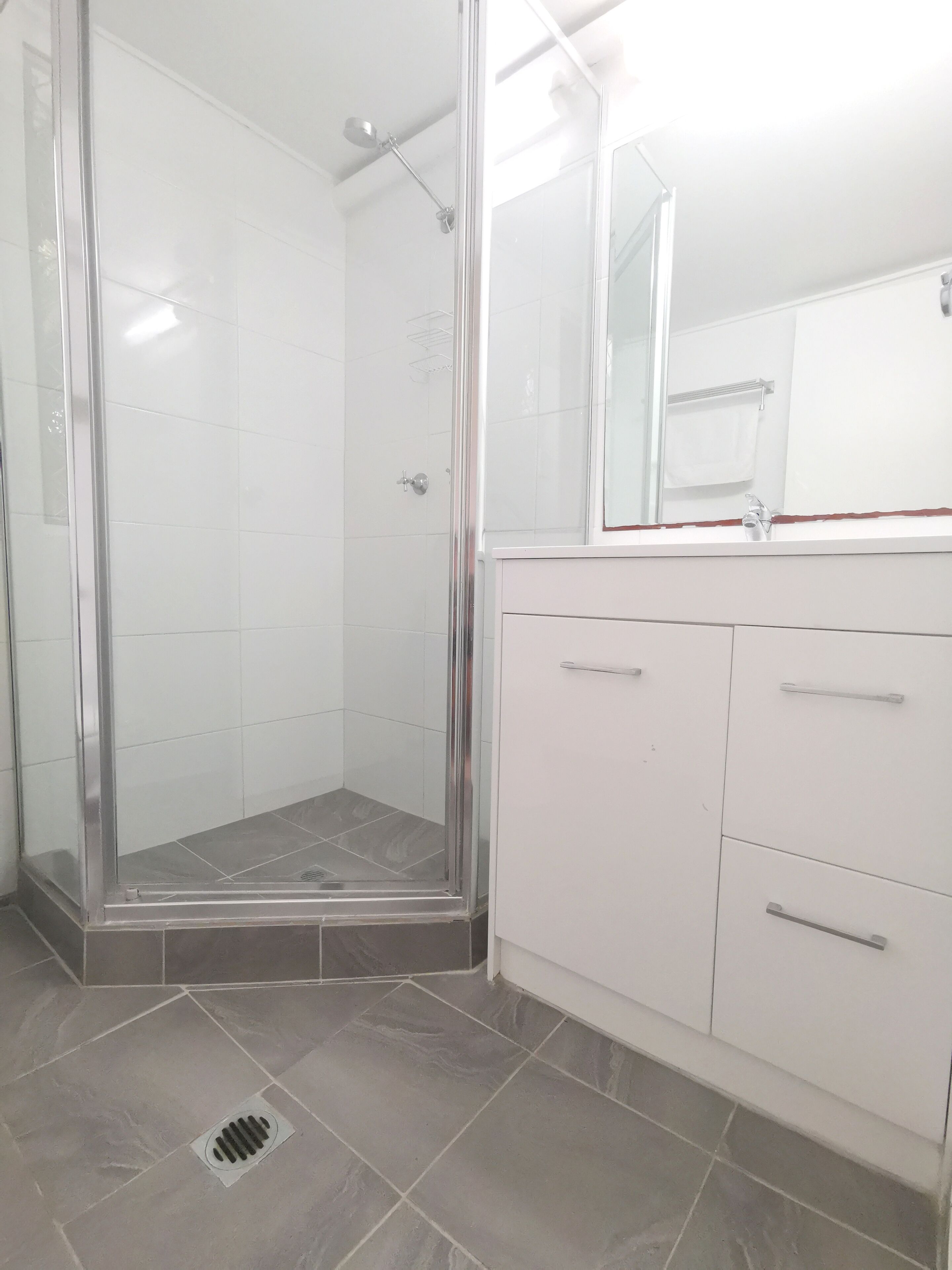 Un baño con ducha de vidrio, paredes con azulejos blancos, tocador blanco con cajones y un espejo sobre el lavabo.