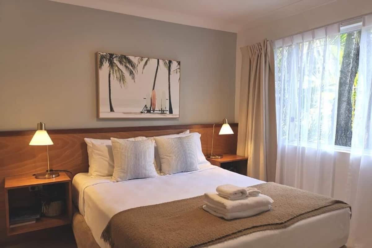 A neatly made bed with white linens and a brown throw blanket, flanked by two bedside lamps and a nightstand with a lamp. A framed picture of palm trees hangs above the bed.