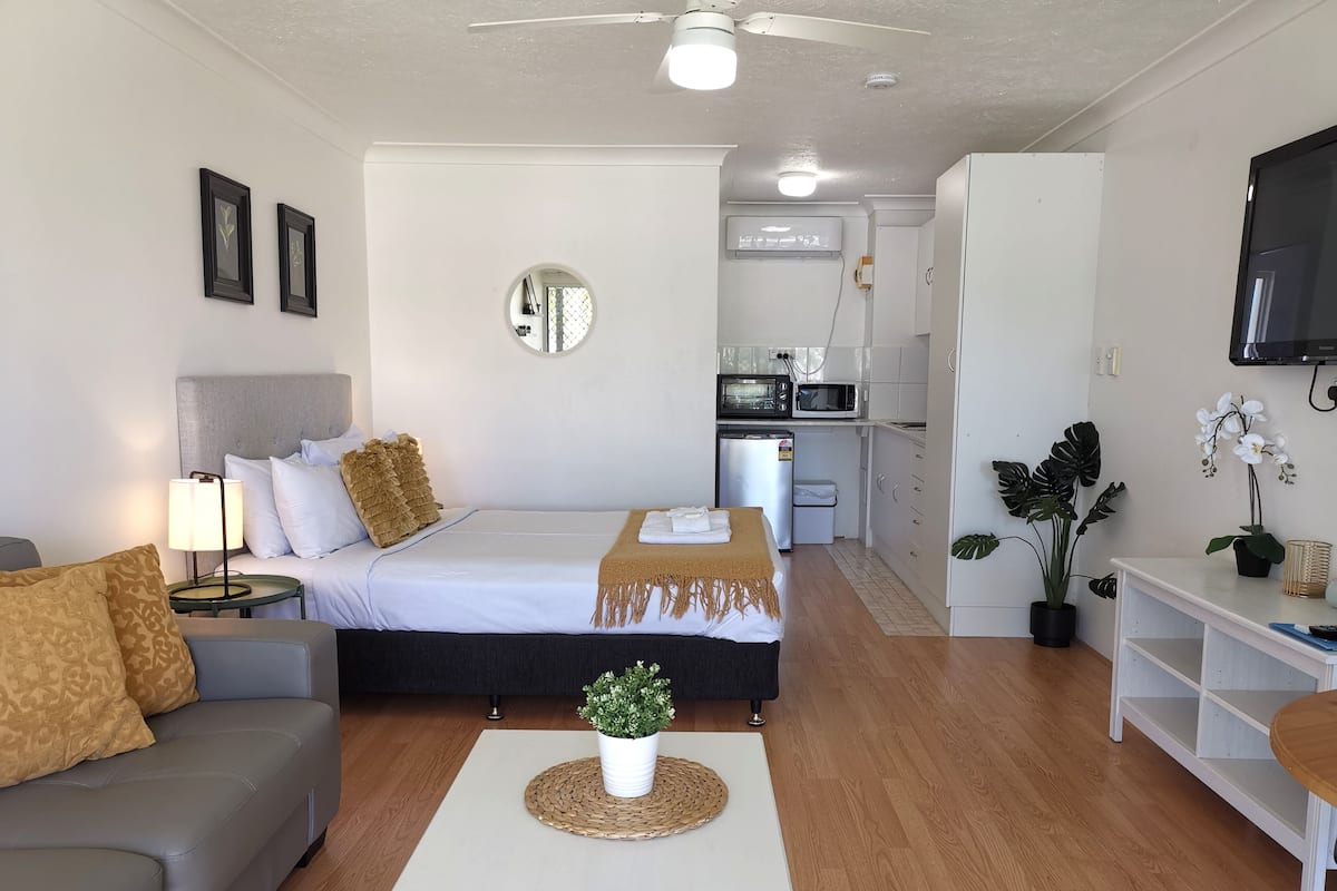 A modern hotel room with a bed, a sofa, a small table with a plant, a kitchen area, and a TV mounted on the wall.