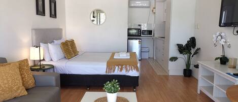 Una habitación de hotel moderna con cama, sofá, una mesita con una planta, zona de cocina y televisor montado en la pared.