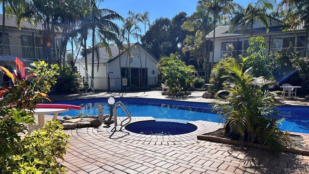 A swimming pool area with a tiled deck, surrounded by palm trees and a small building. - Bay of Palms (Elanora)