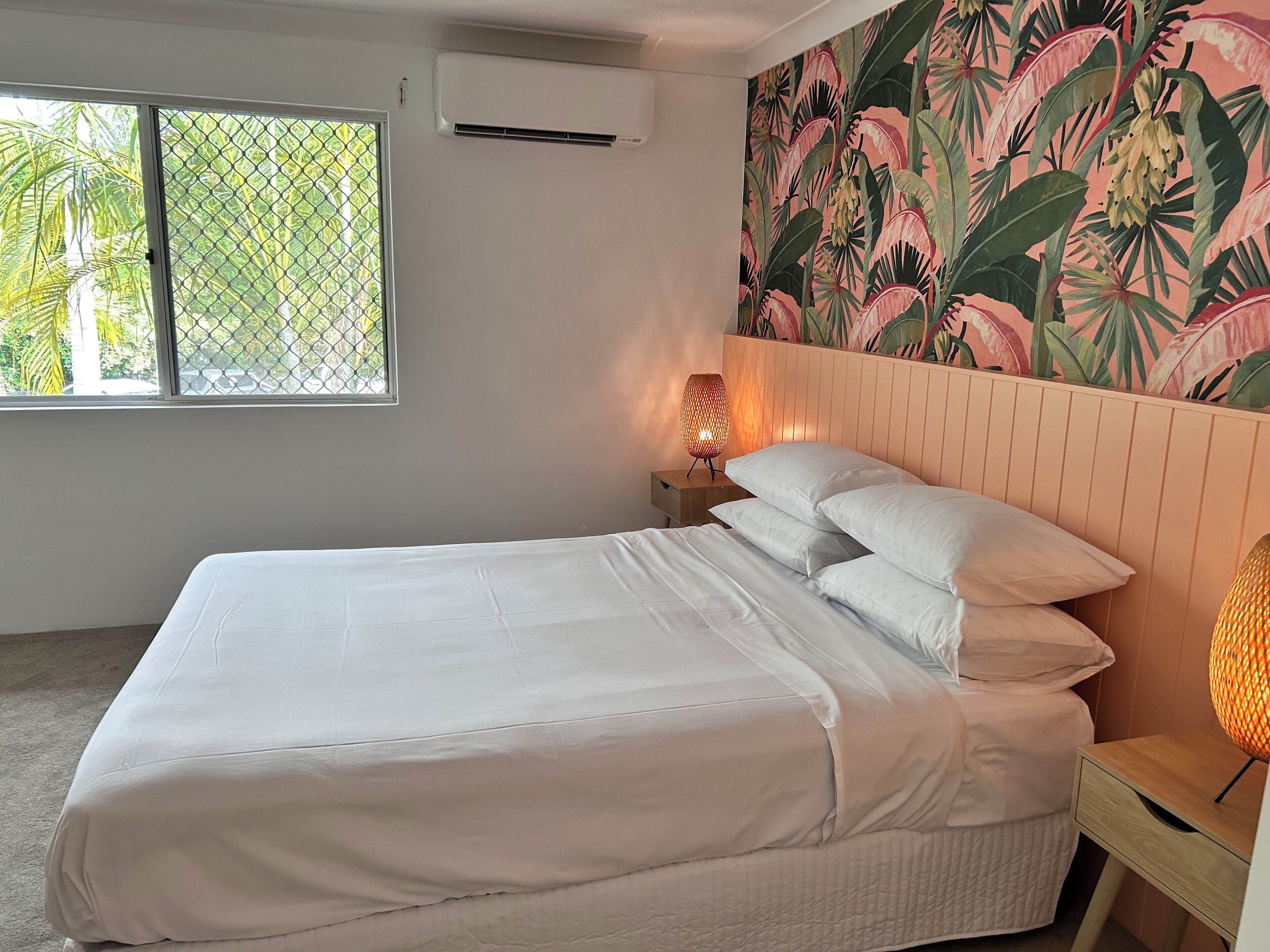 A neatly made bed with white linens, a wooden nightstand with a lamp, and a tropical-themed wall mural.