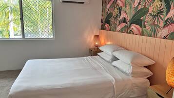 A neatly made bed with white linens, a wooden nightstand with a lamp, and a tropical-themed wall mural.