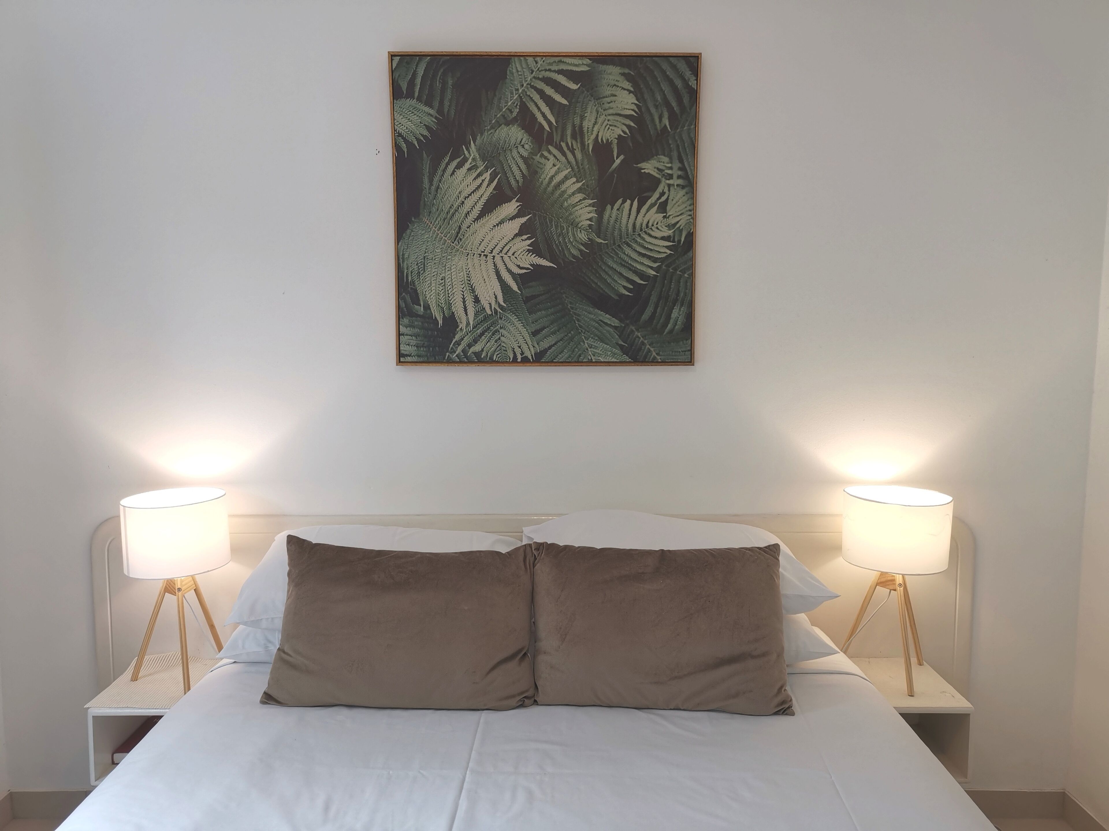 Un letto rifatto con due cuscini, affiancato da comodini con lampade, e un'opera d'arte con una foglia di felce al muro.
