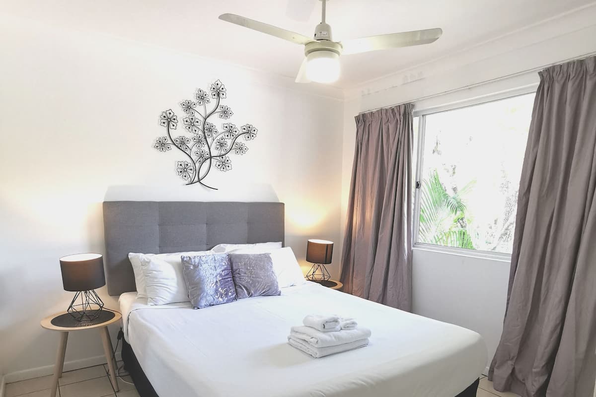 A neatly made bed with a white comforter and folded towels, a grey headboard, a wall-mounted tree decoration, a ceiling fan, and a window with curtains.