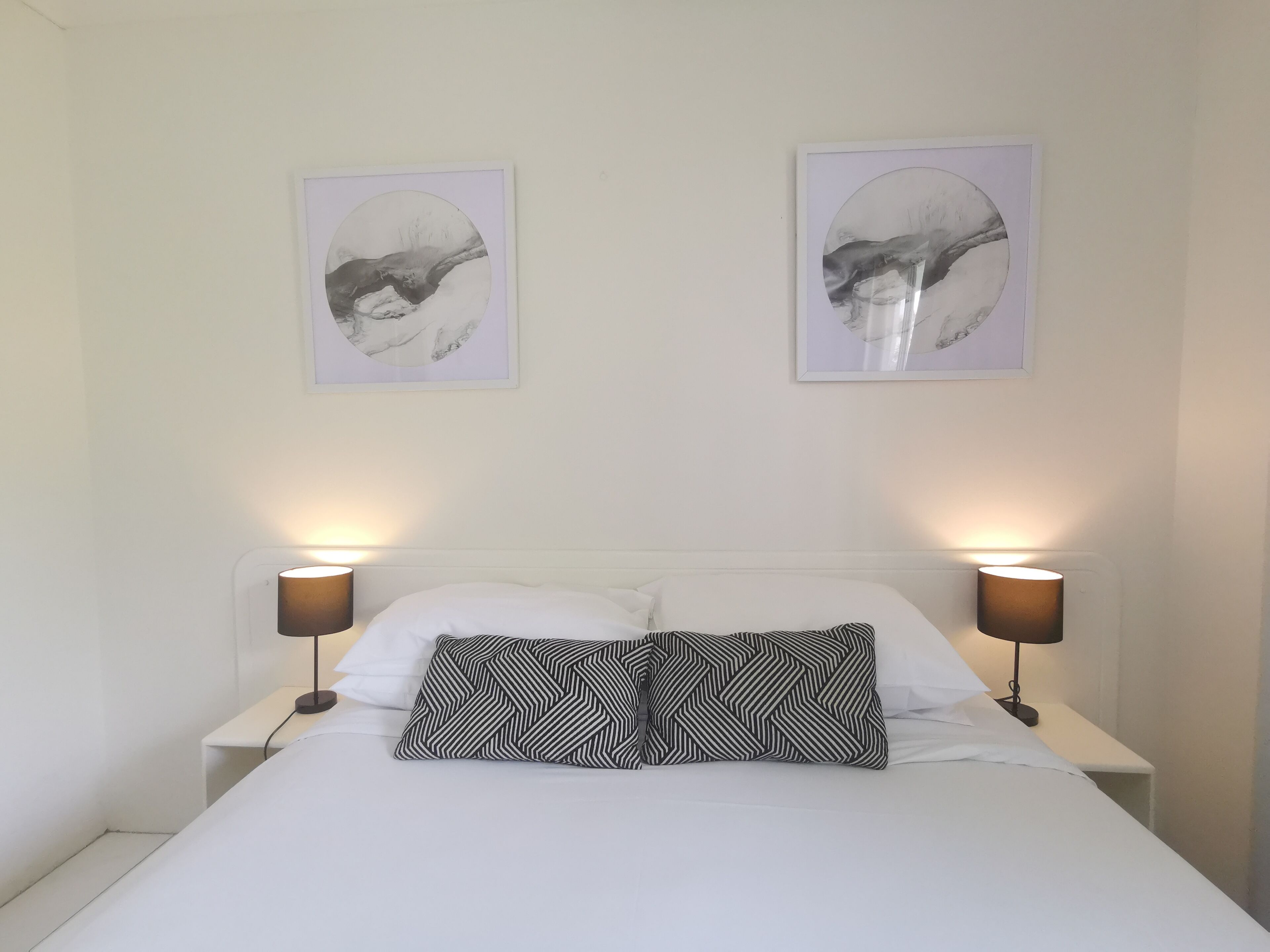 Cama bem arrumada com duas almofadas, duas luminárias fixadas na parede e dois quadros enquadrados.