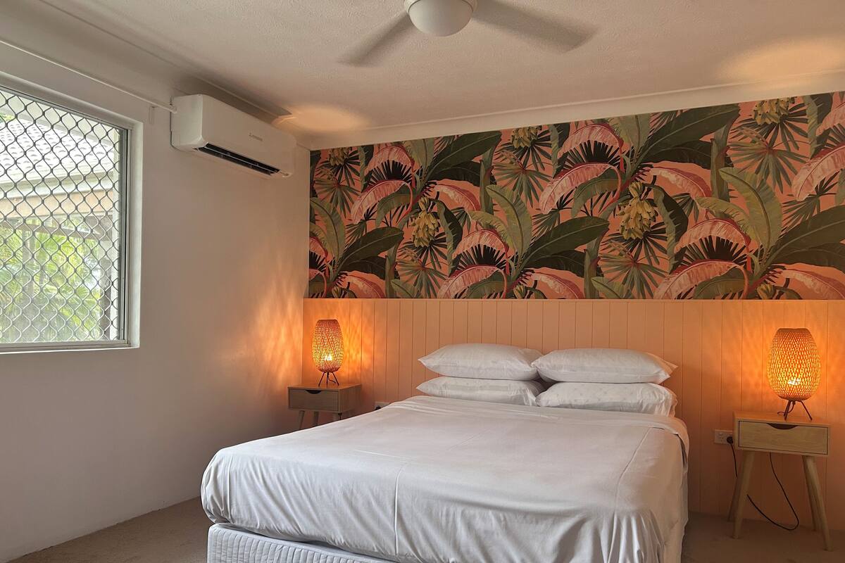 A bedroom with a bed, two bedside lamps, a window with a mesh screen, and a tropical leaf-patterned wall.