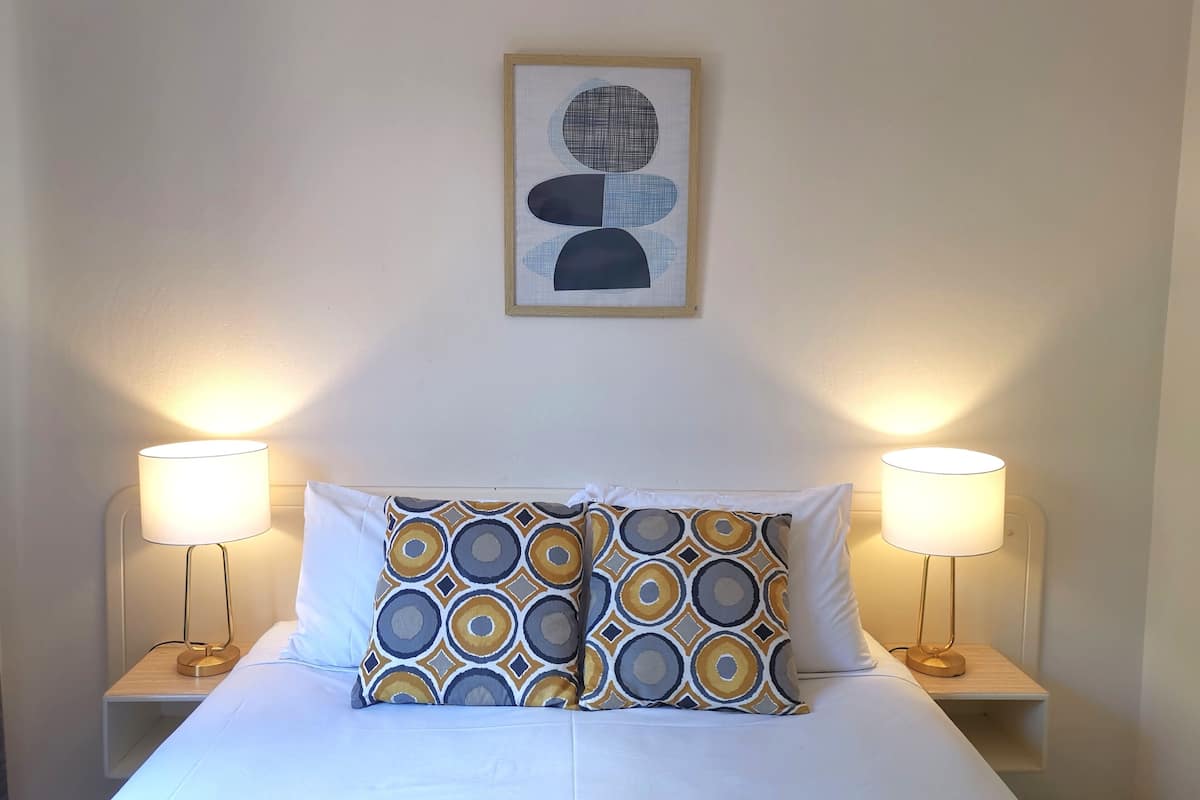 A neatly made bed with patterned pillows, two bedside lamps, and a framed abstract artwork on the wall.