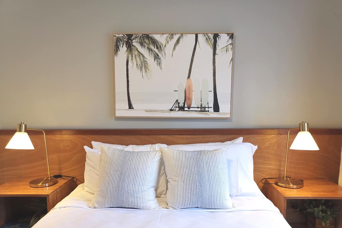 A neatly made bed with two pillows, flanked by two bedside tables with lamps, and a framed picture of palm trees on the wall.