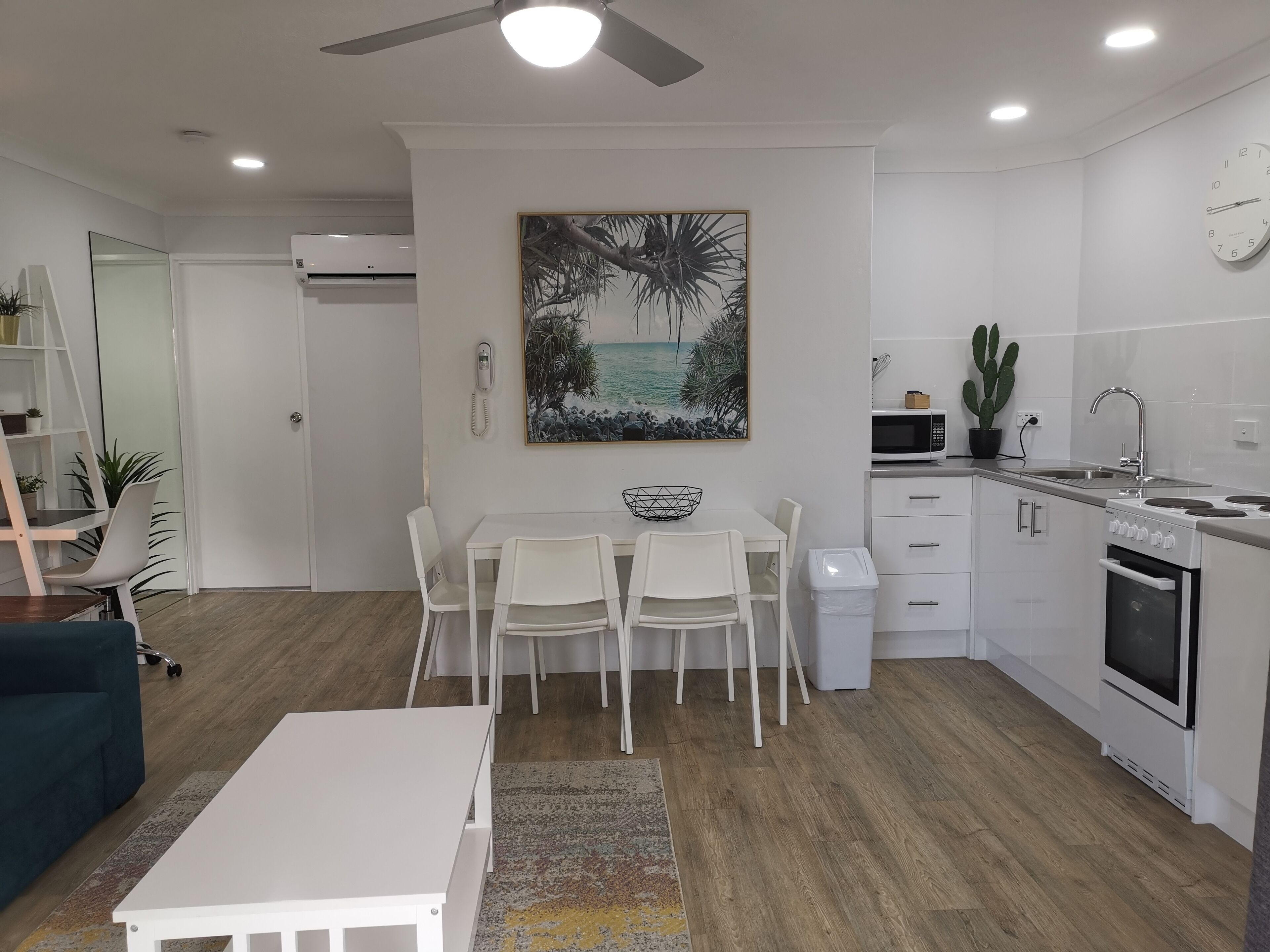 Une cuisine moderne avec des meubles blancs, un coin repas comprenant une table et des chaises blanches, un ventilateur de plafond, un tableau représentant une scène de plage et une horloge au mur.