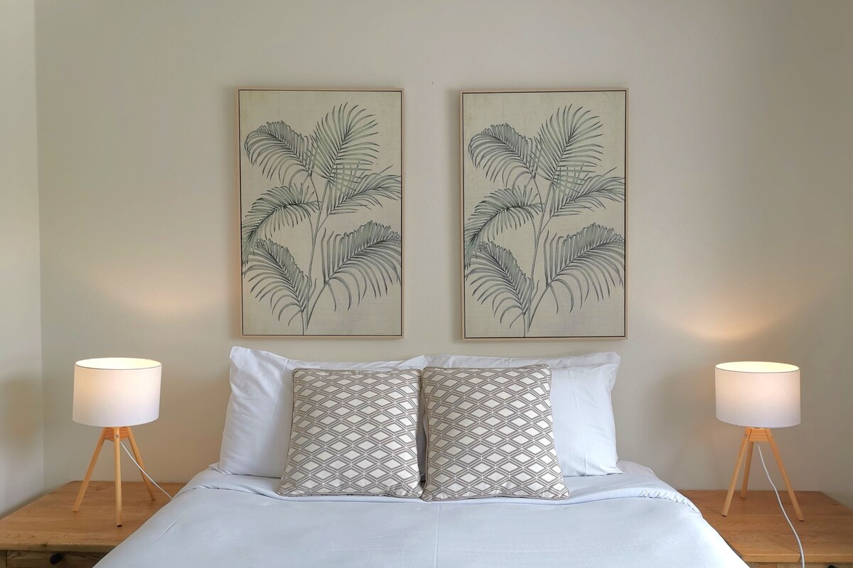 A bedroom with a bed, two patterned pillows, a nightstand, a lamp, and two framed botanical prints on the wall.