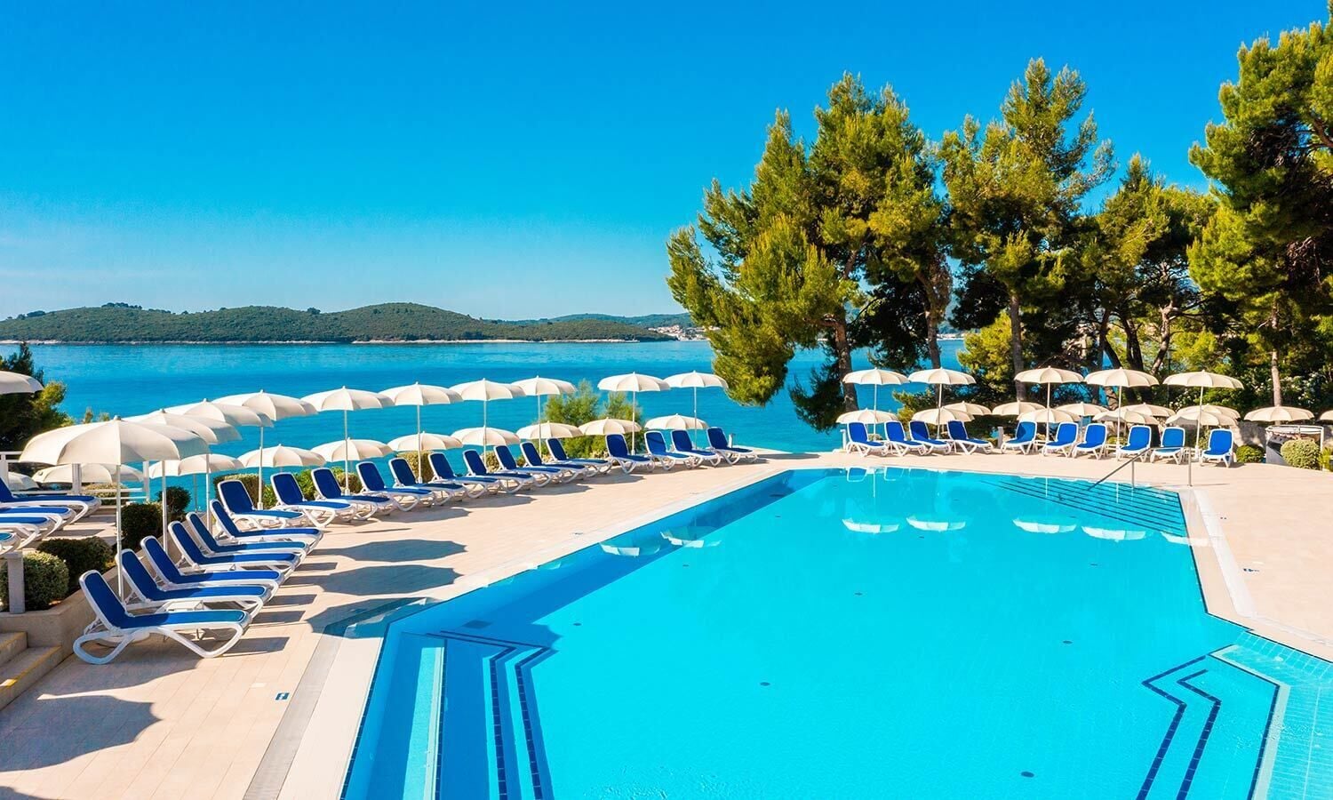 A swimming pool with blue water, surrounded by lounge chairs and umbrellas, with a view of the sea and hills in the background.