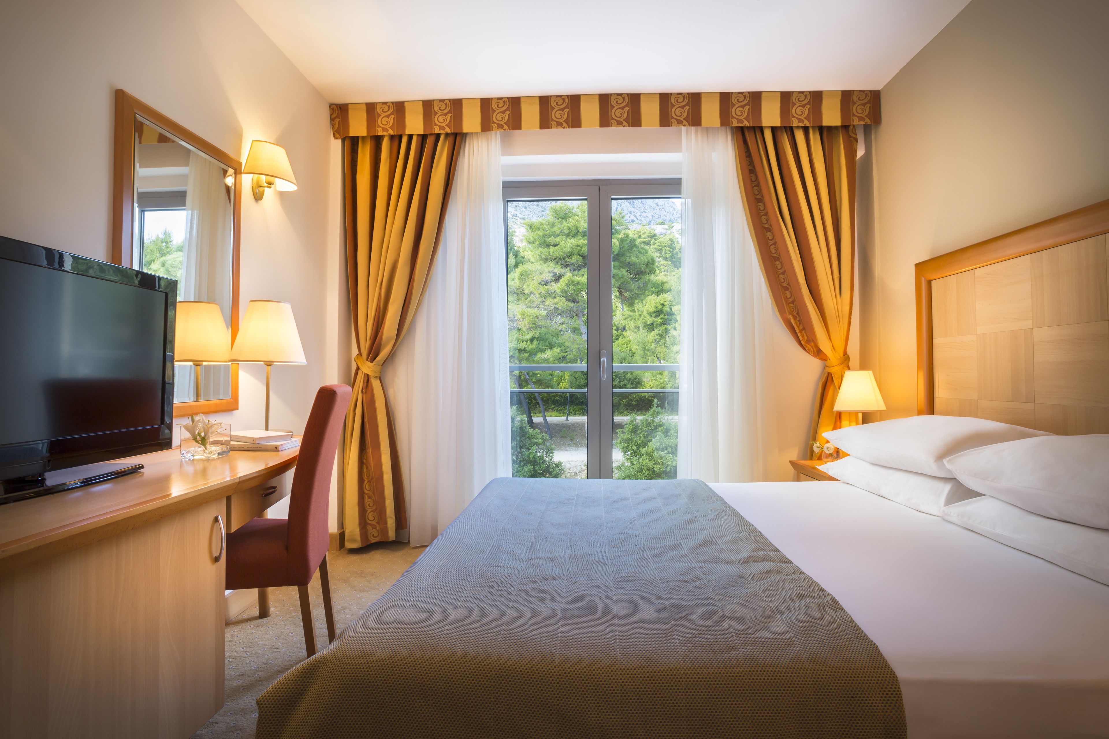 Habitación de hotel con cama, escritorio con televisor, dos lámparas y vista a zonas verdes por la ventana.