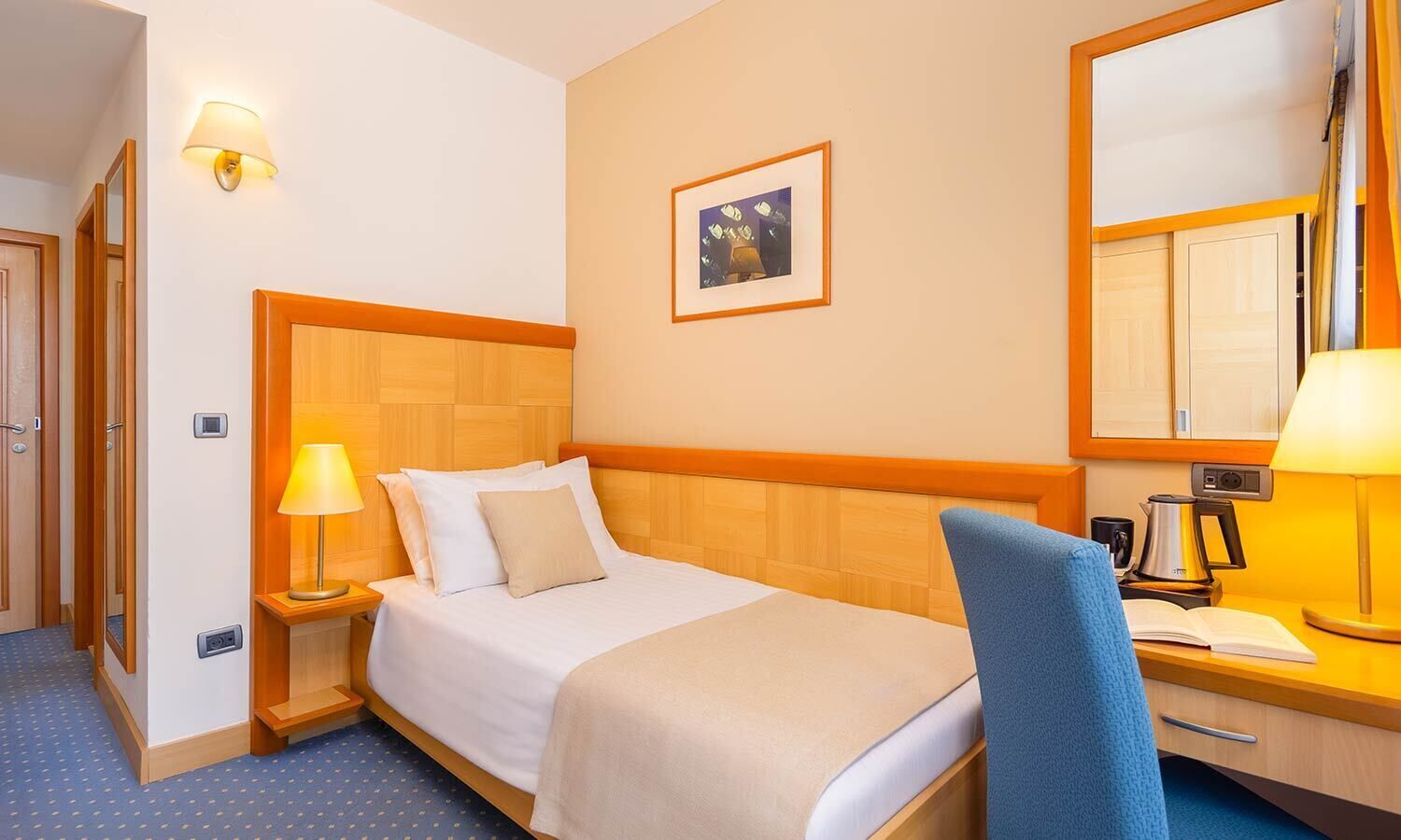 Habitación de hotel con cabecera de madera, cama con sábanas blancas, mesita de noche con lámpara, una silla y escritorio con lámpara y un libro.