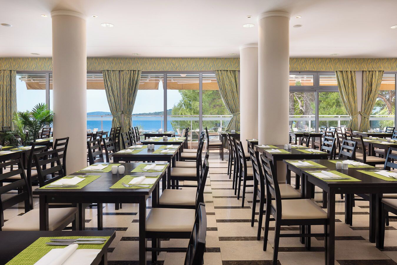 Een restaurant met gedekte tafels, grote ramen met gordijnen en uitzicht op zee.