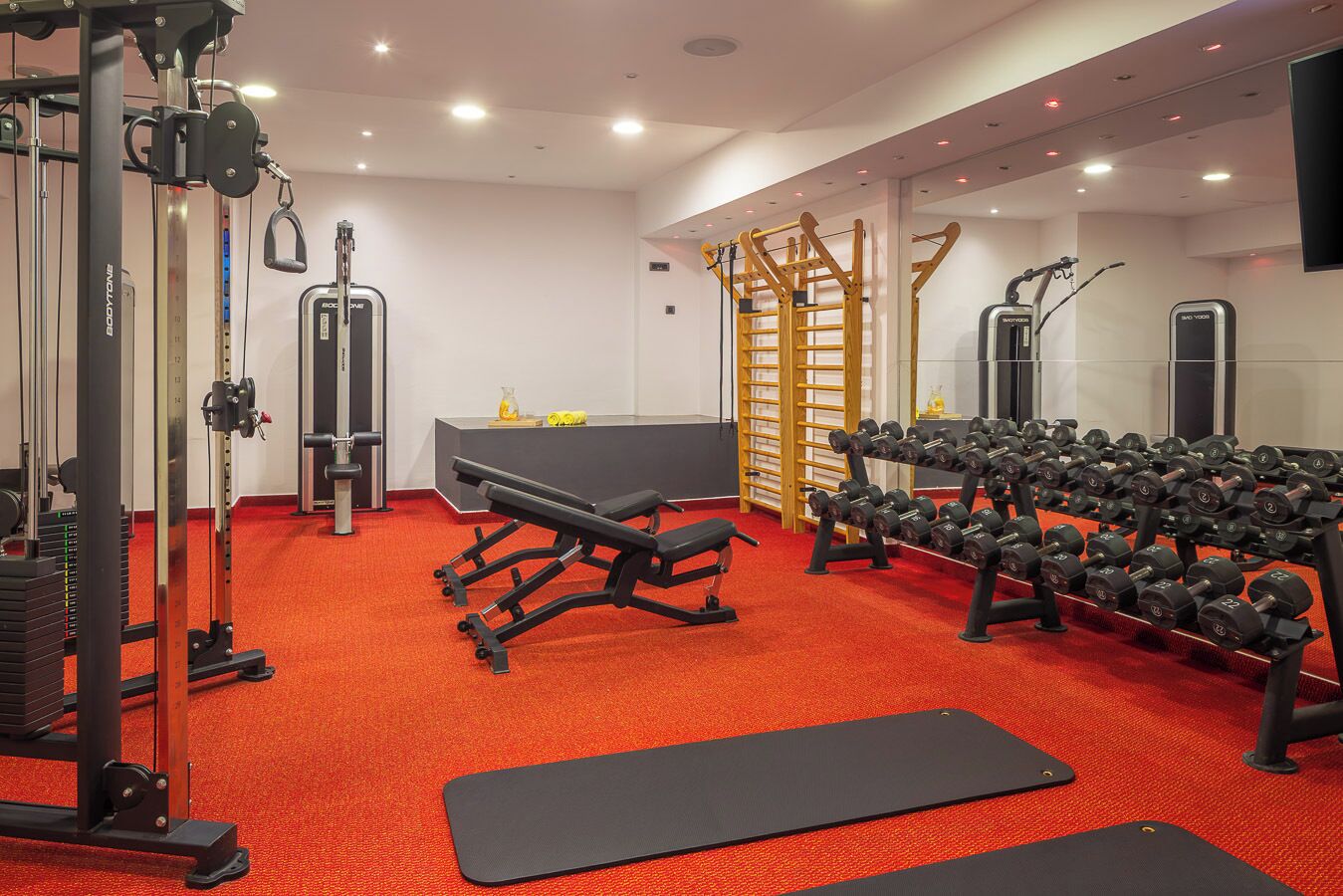 A gym with exercise equipment, a bench, dumbbells, and a red carpet floor.