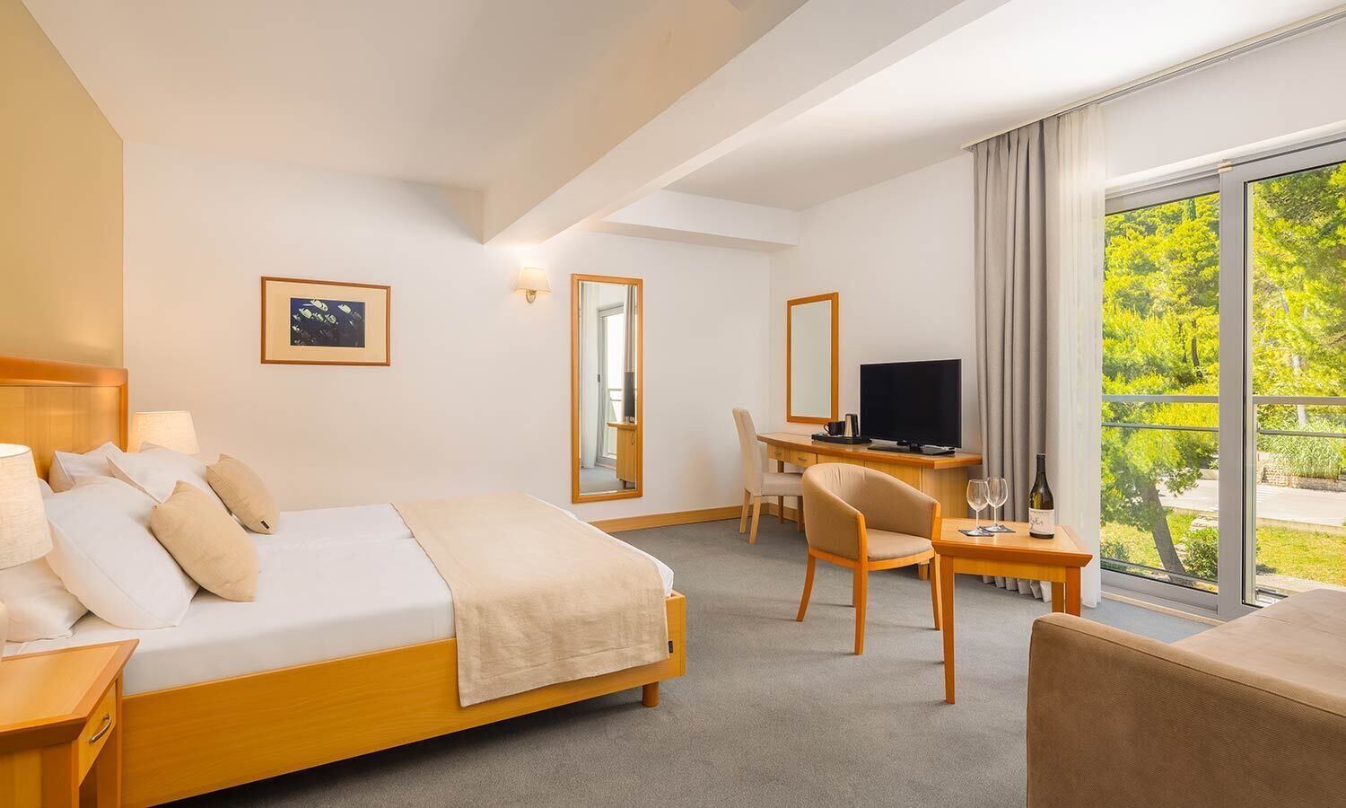 A hotel room with a bed, a desk, a chair, a television, and a window with a view of greenery.