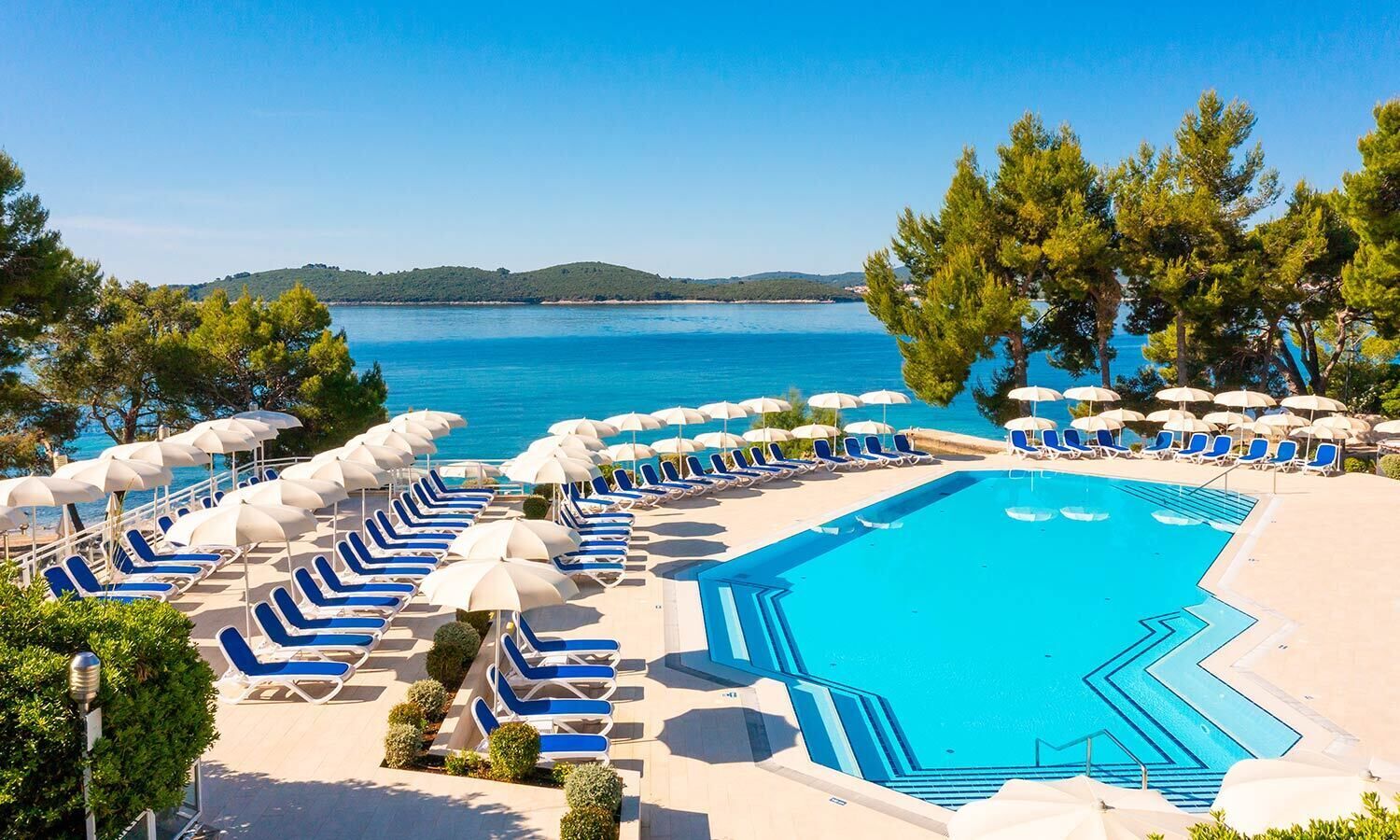 A poolside area with blue lounge chairs and umbrellas, a clear blue pool, and a view of the sea and hills in the background.