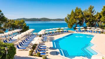 A poolside area with blue lounge chairs and umbrellas, a clear blue pool, and a view of the sea and hills in the background.