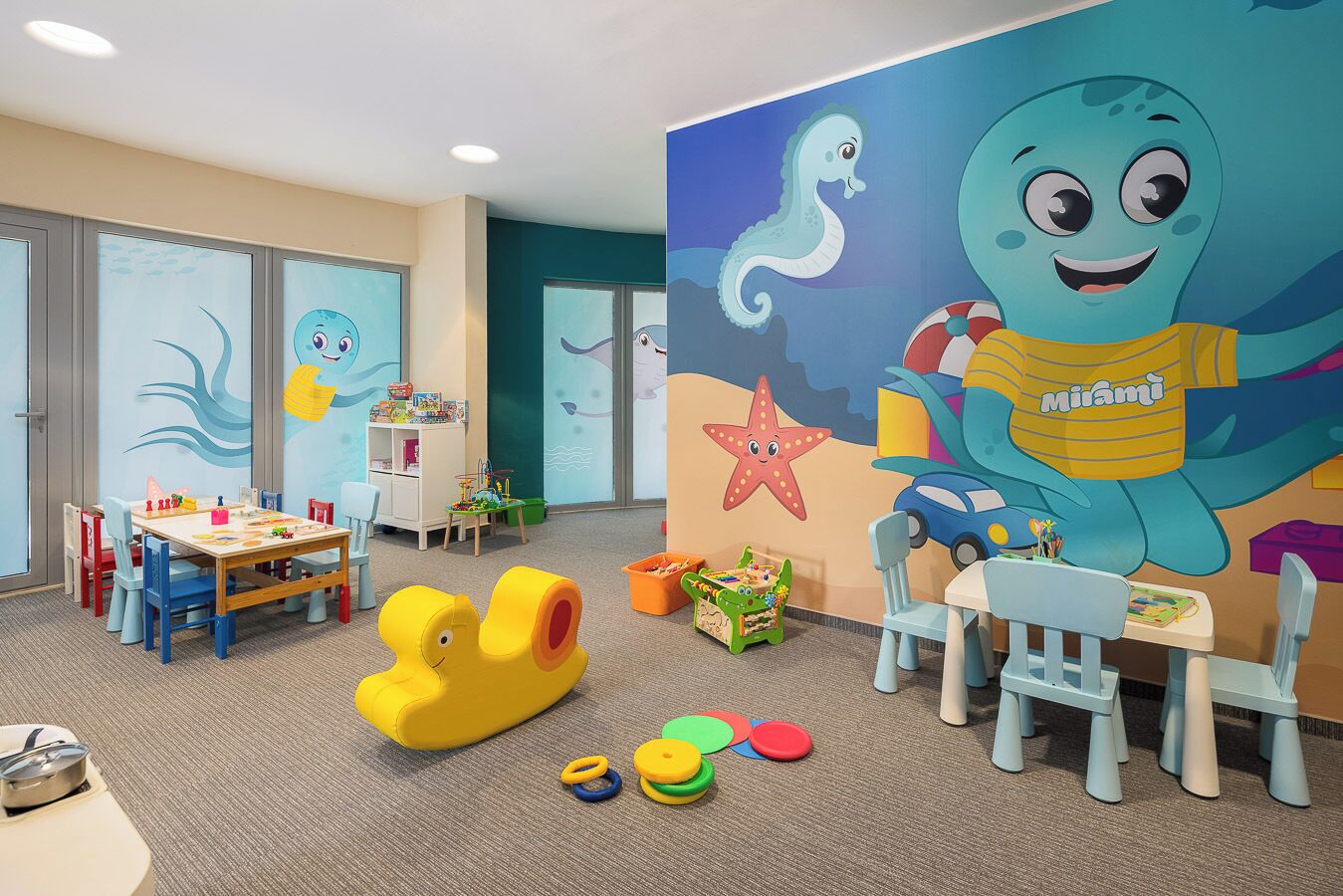 Un salón de juegos infantiles con un mural grande, mesas de juego y juguetes coloridos.