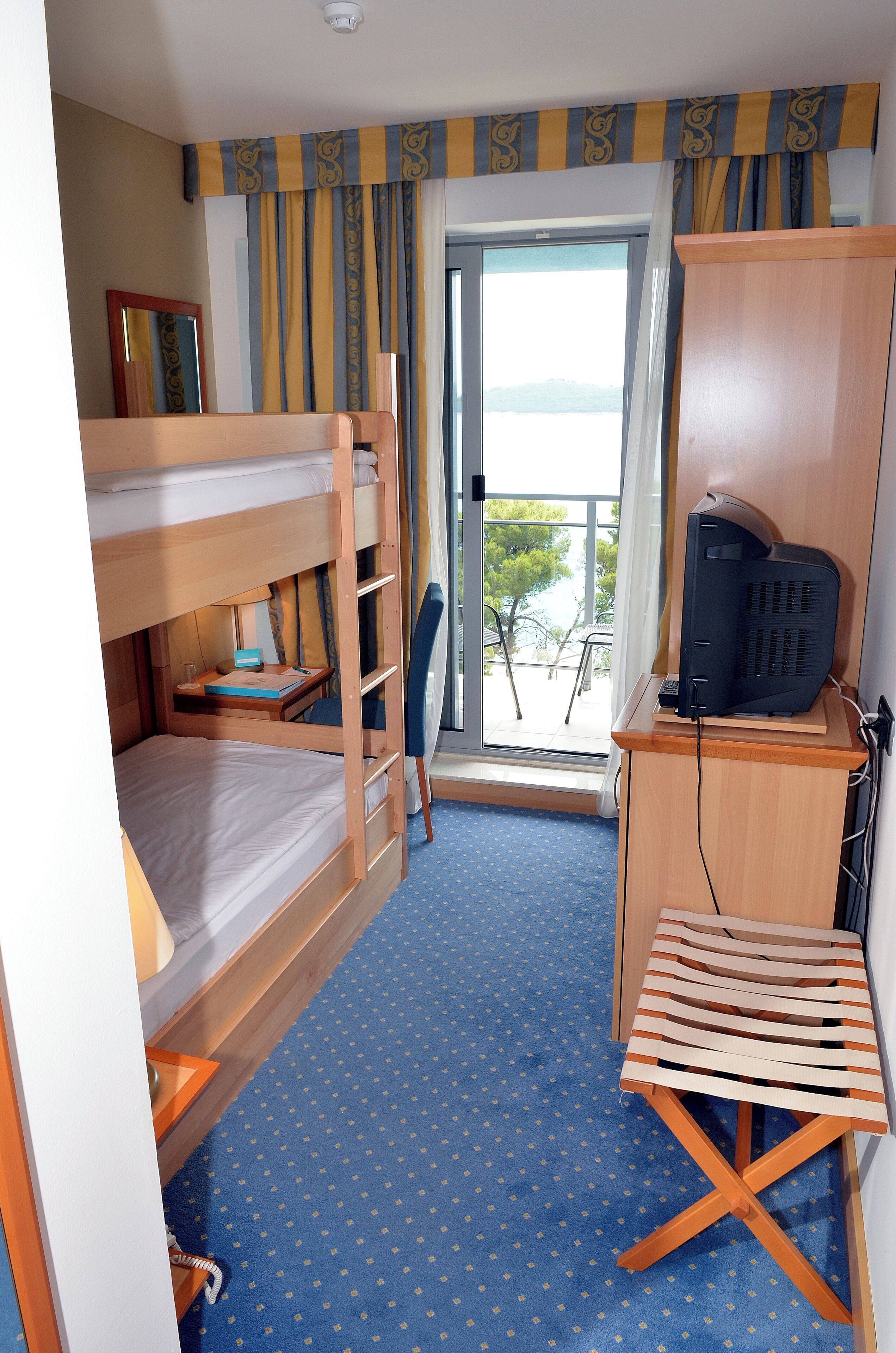 Ein kleines Hotelzimmer mit Etagenbett, Schreibtisch inklusive Computer, Stuhl und Blick ins Freie.