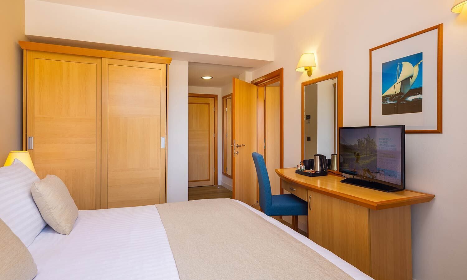 Ein Hotelzimmer mit Bett, Schreibtisch mit Computer, Stuhl, Kleiderschrank und einem gerahmten Bild an der Wand.