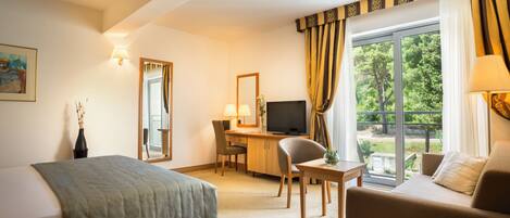 A hotel room with a bed, a desk with a computer, a TV, a sofa, and a window with curtains.