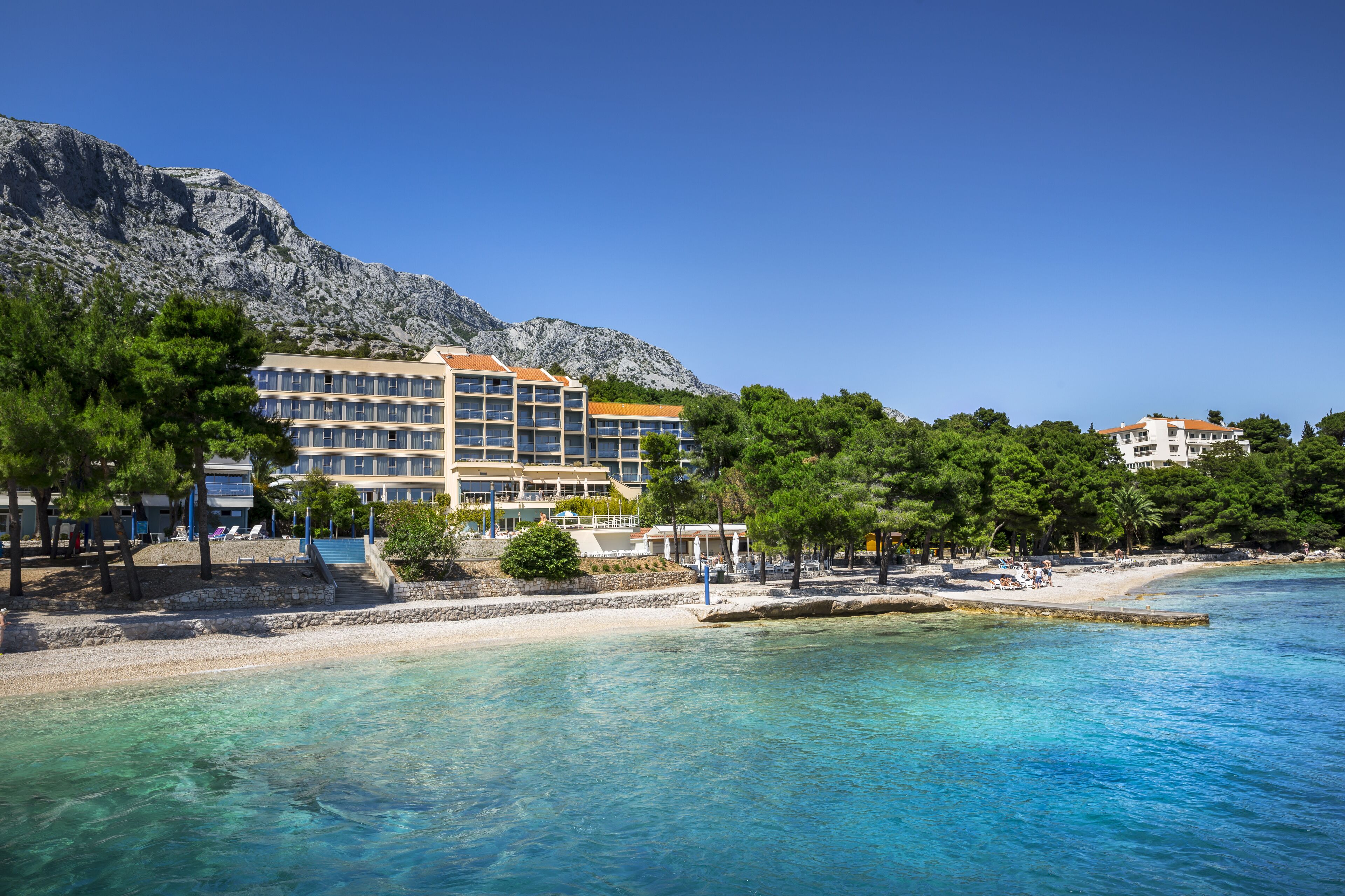 A coastal hotel with multiple floors, a beach, and a mountainous backdrop.