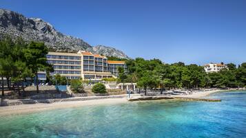 A coastal hotel with multiple floors, a beach, and a mountainous backdrop.