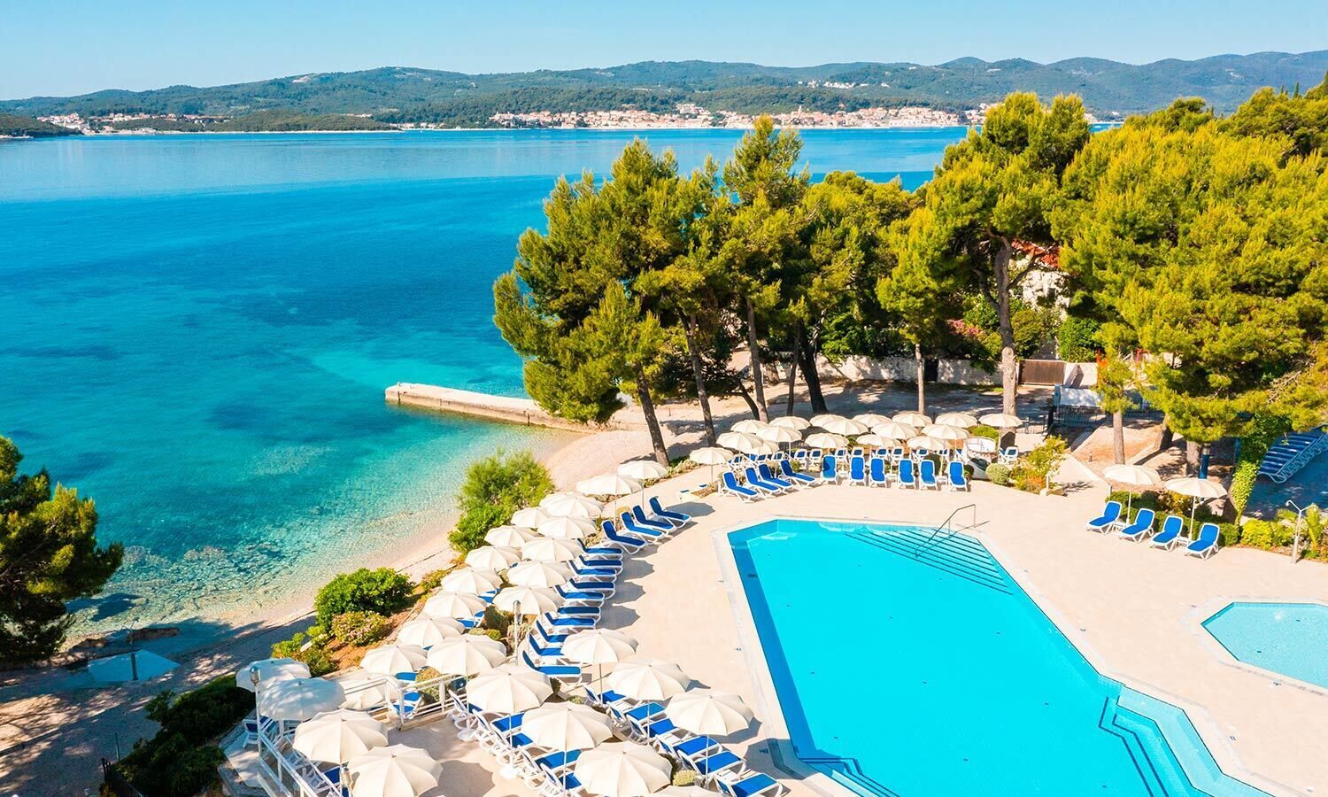 A resort pool area with clear blue water, lounge chairs, and umbrellas, surrounded by trees and a view of the sea.